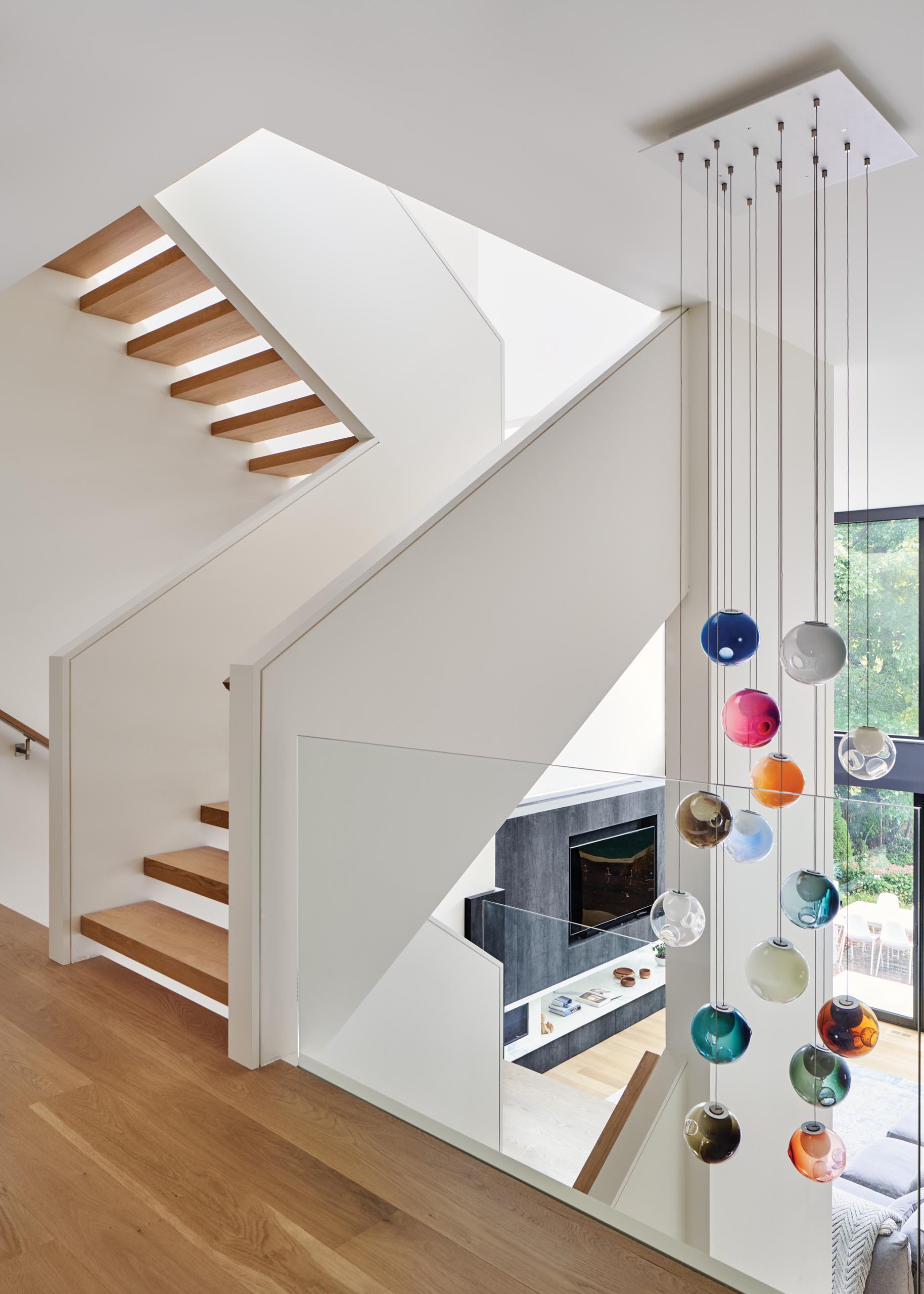 A modern house with an open wood staircase that has glass railings that connect the multiple levels of the home. A colorful accent has been added with the inclusion of suspended Bocci lights.