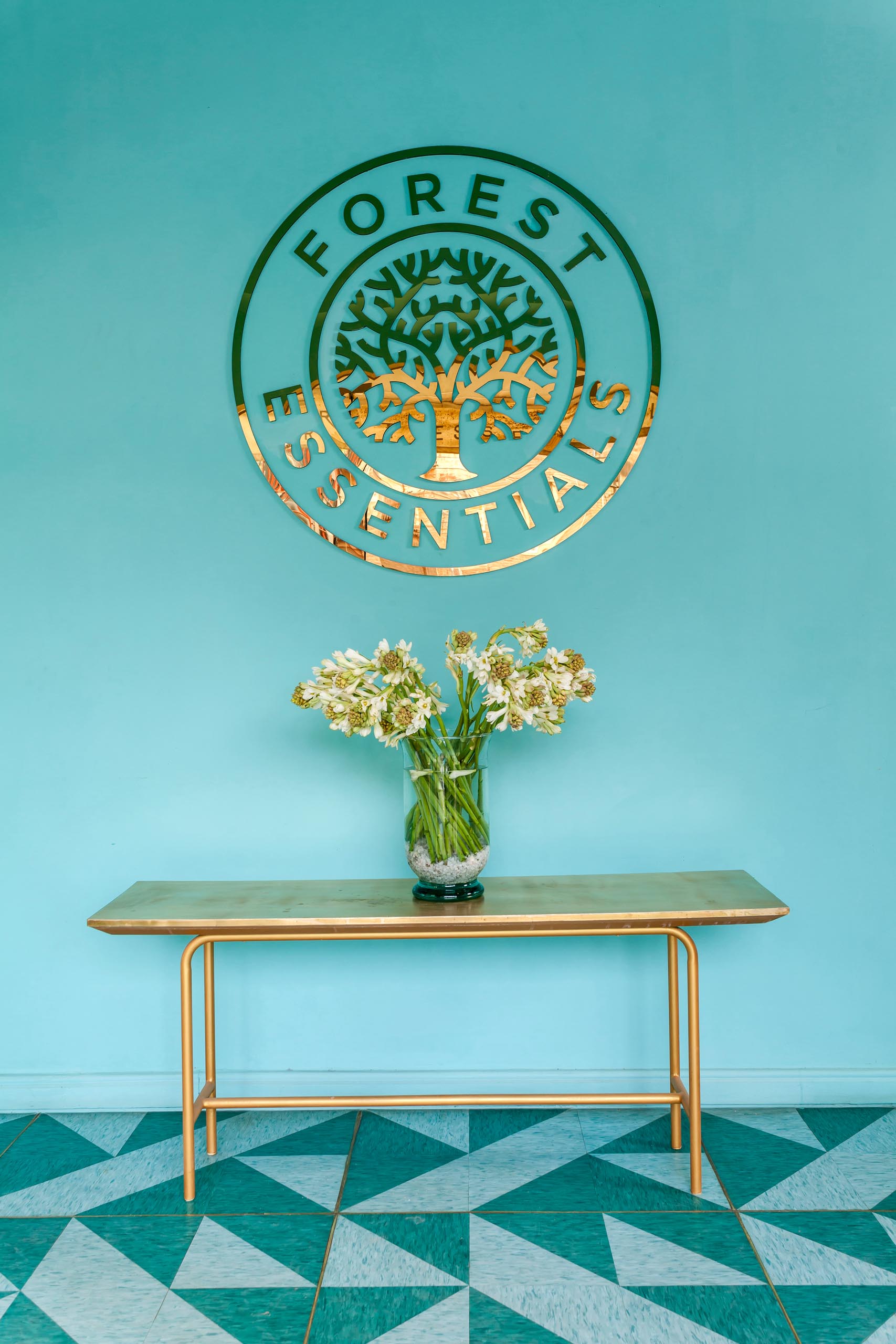 A modern retail store entryway with teal walls, a gold logo, a gold bench, and patterned floor.