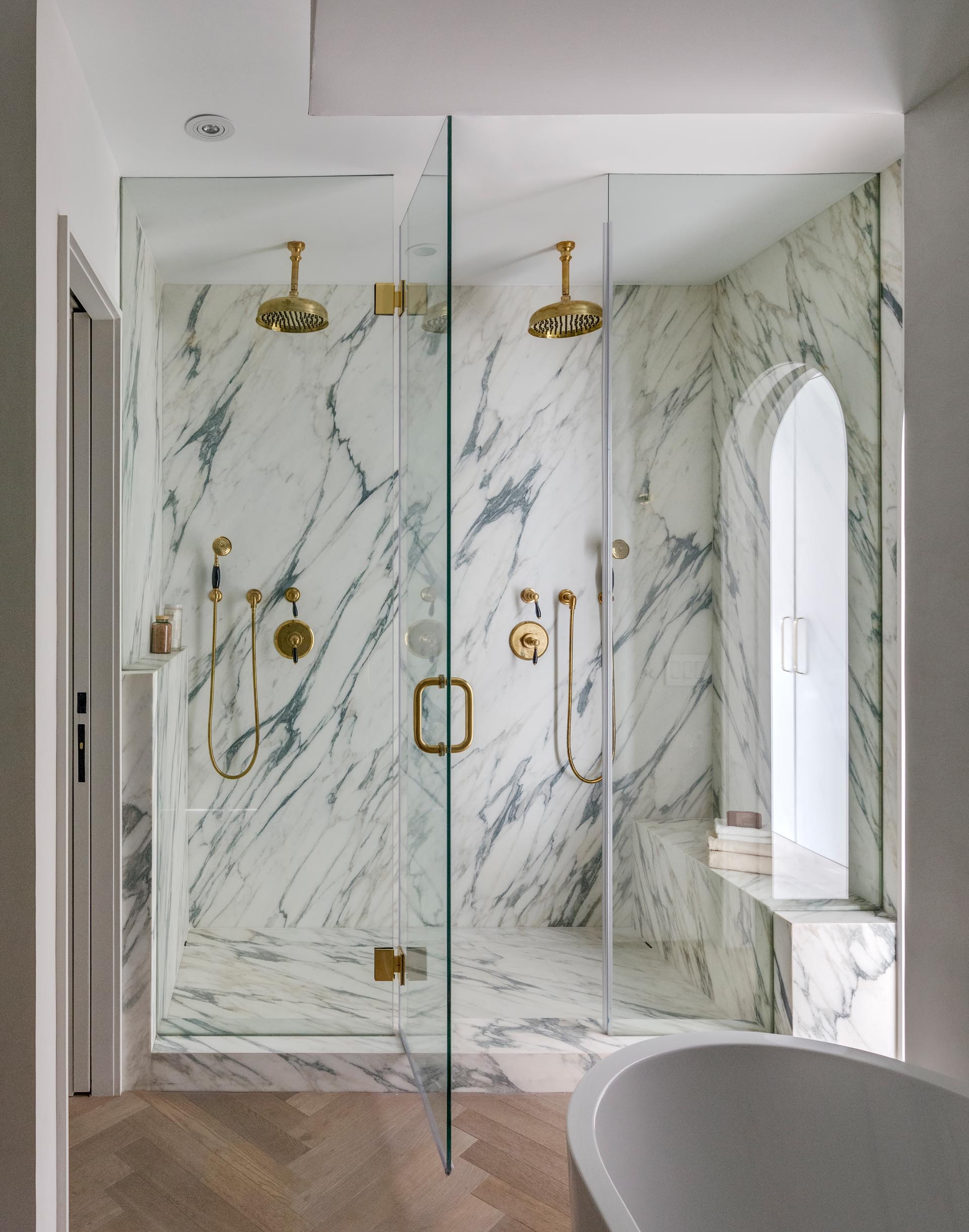 A modern walk-in shower with gold hardware, two shower heads, a glass shower screen and door, and a built-in bench.