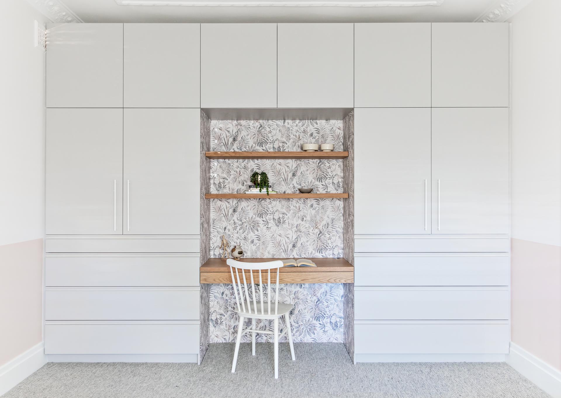 A bedroom with a wall of cabinets and drawers that surround a built-in wood desk and shelving. The desk nook is also lined with a botanical wallpaper.