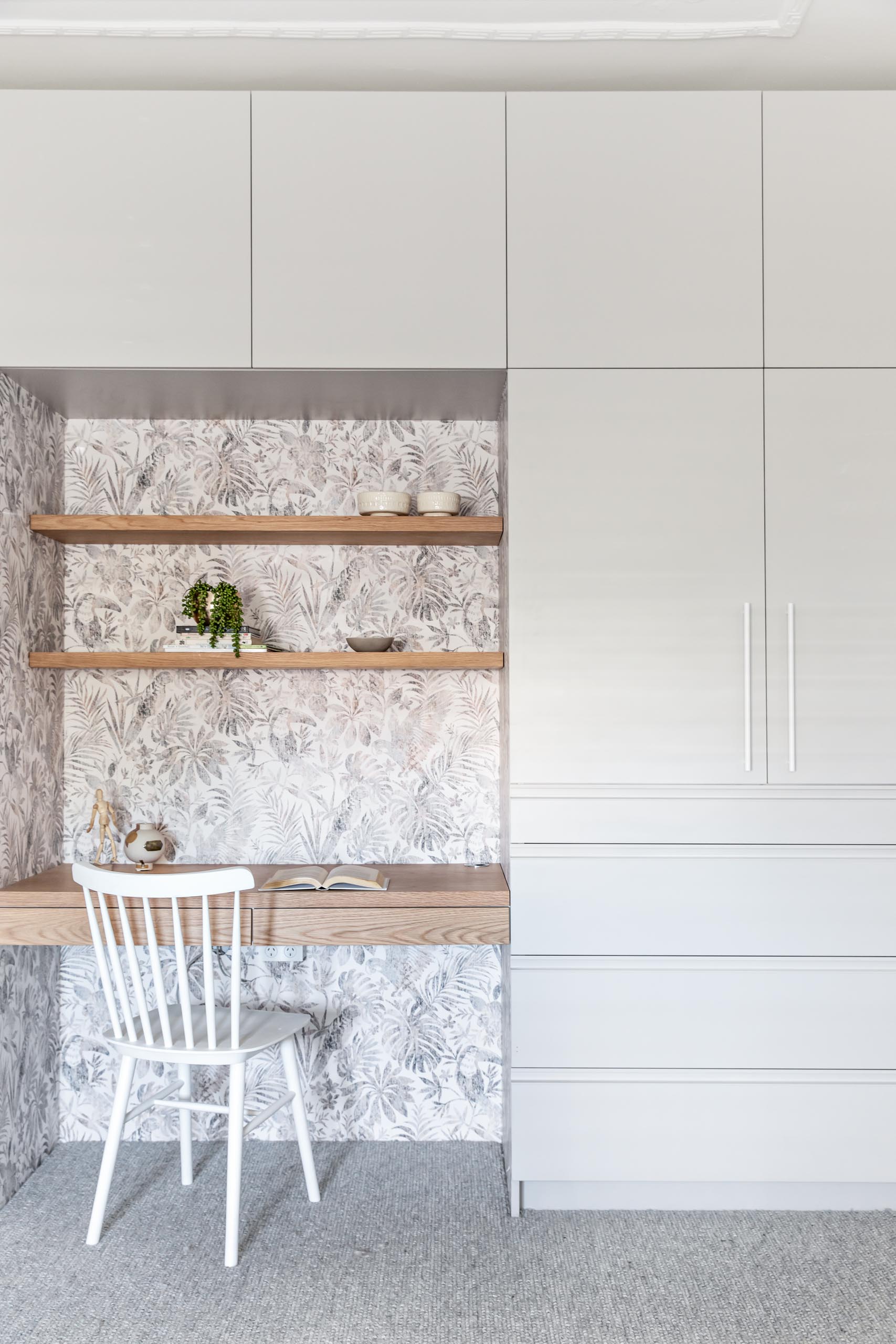 A wall of cabinets and drawers surround a built-in wood desk and shelving. The desk nook is also lined with a botanical wallpaper.