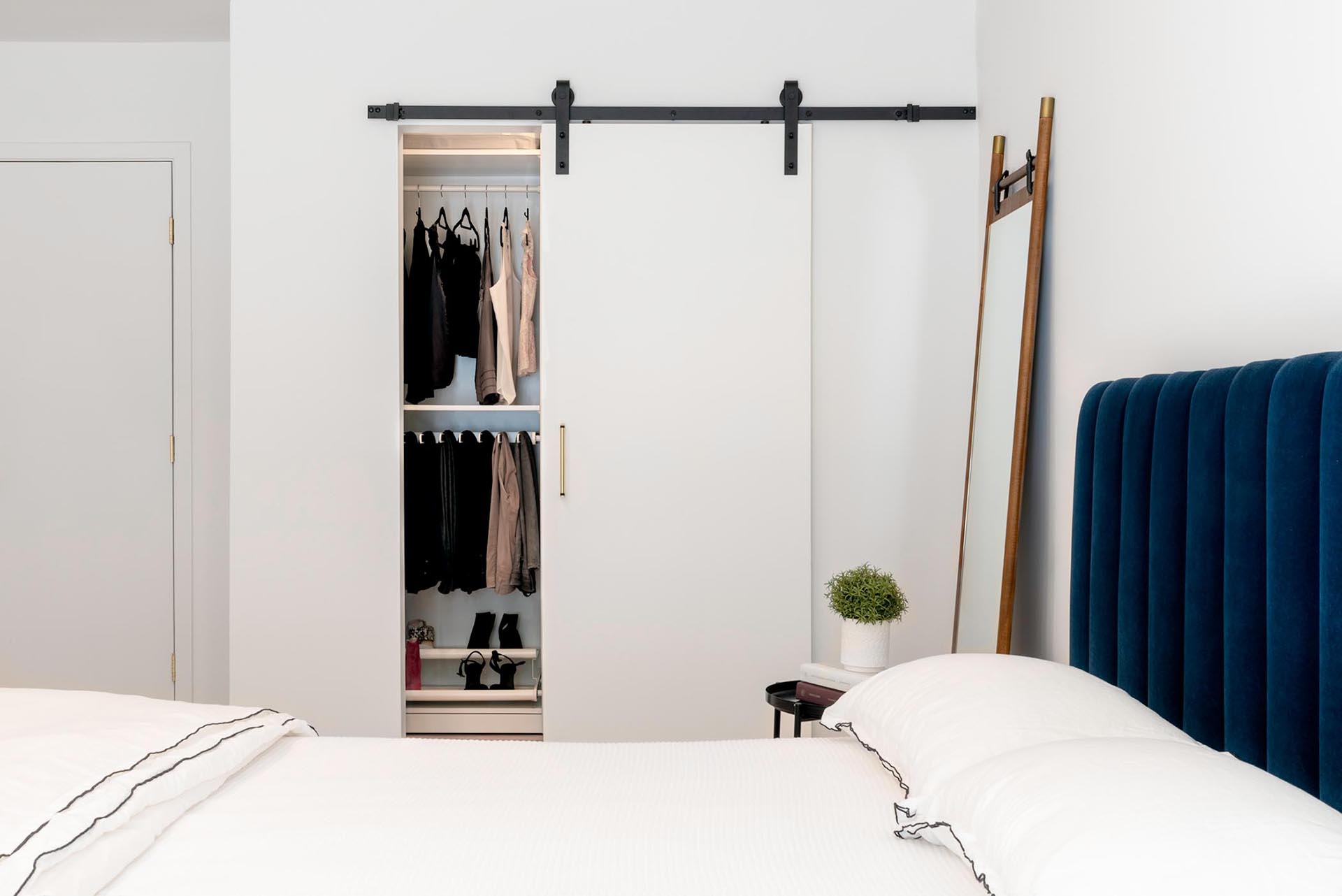 In this modern bedroom, there's a sliding barn door on the closet that includes matte black hardware. Color has been added to the interior with the use of a deep, royal blue velvet headboard and bed frame.