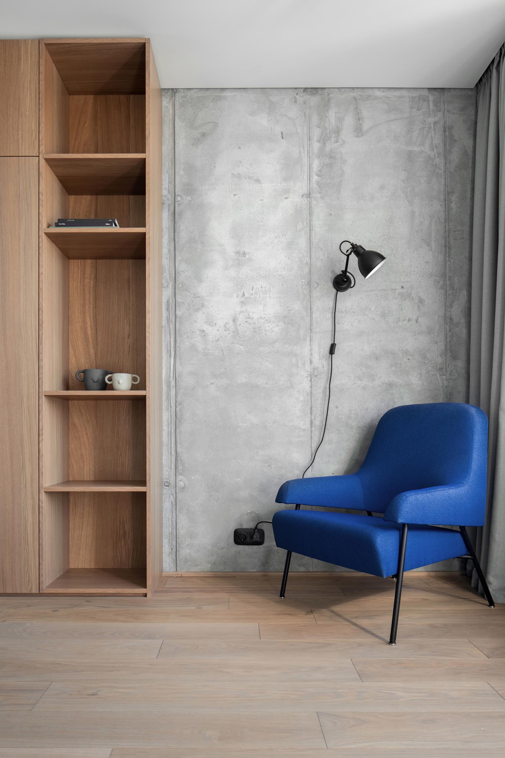 A modern home office with long curtains, a blue armchair, wood shelving, and a concrete wall.