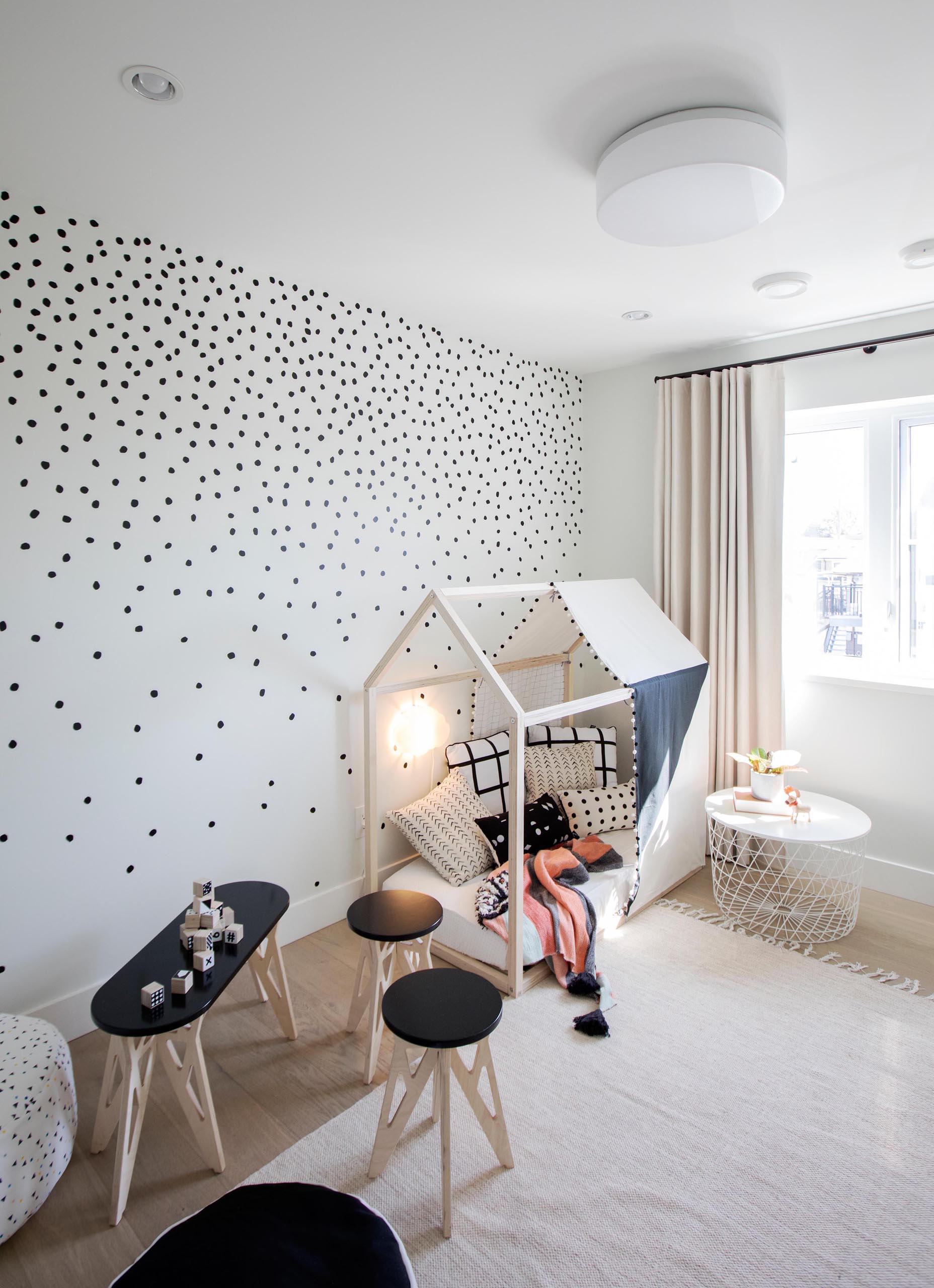 A modern kids bedroom has a playful appearance, with a polka dot pattern on the walls, a small tent, and kid-sized furniture with a table, stools, and poufs.