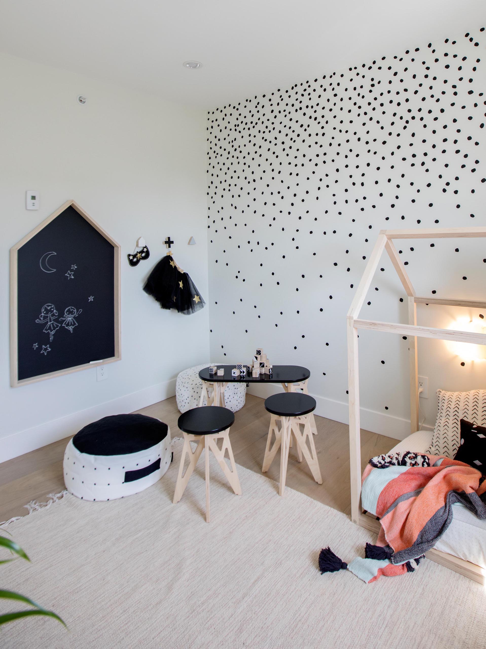 A modern kids bedroom has a playful appearance, with a polka dot pattern on the walls, a small tent, and kid-sized furniture with a table, stools, and poufs.