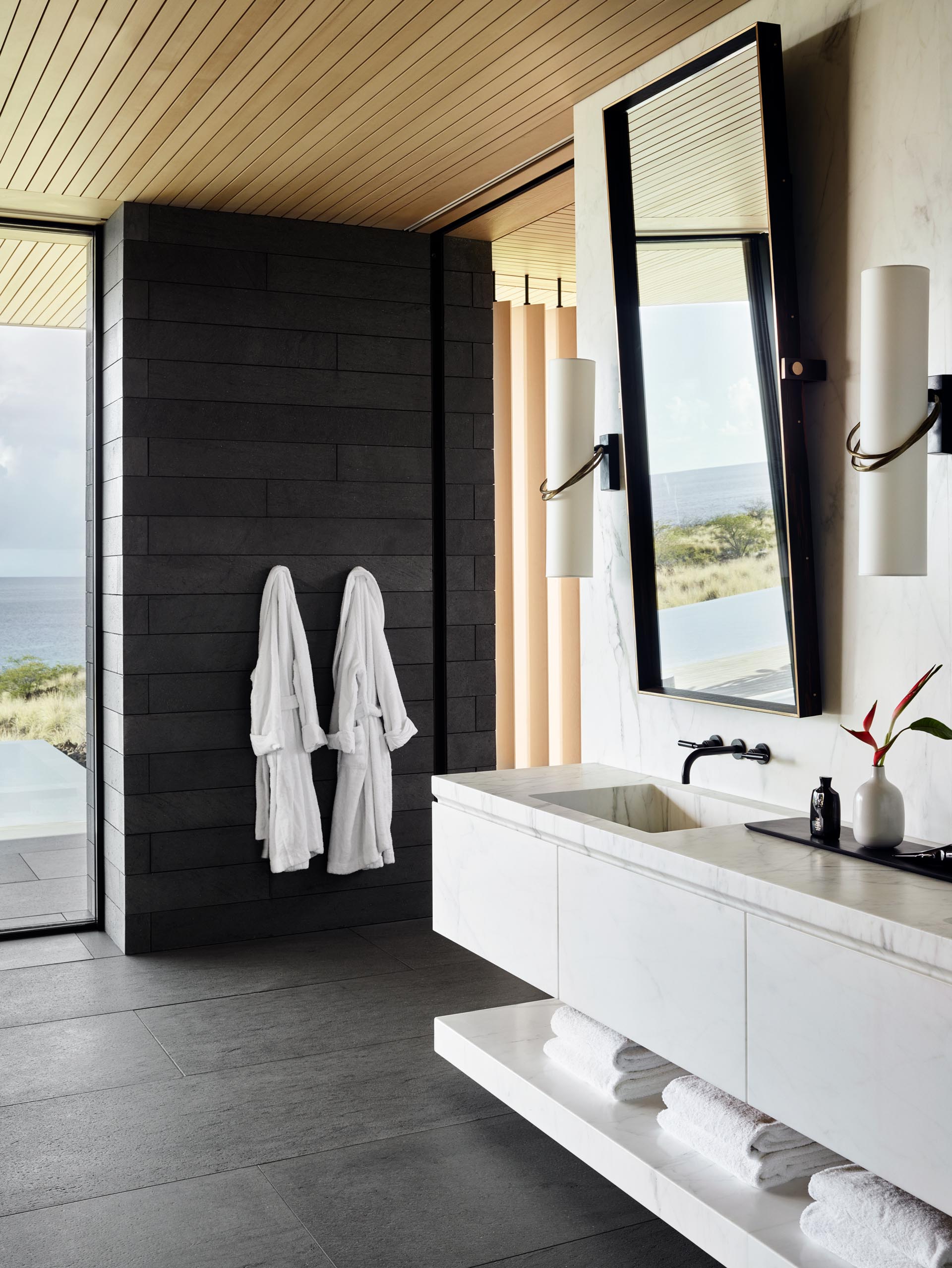 In the master bathroom, large format gray tiles cover the floor, vertical wood screens create privacy, and a white double-sink vanity floats above the ground and is adjacent to the glass-enclosed shower.