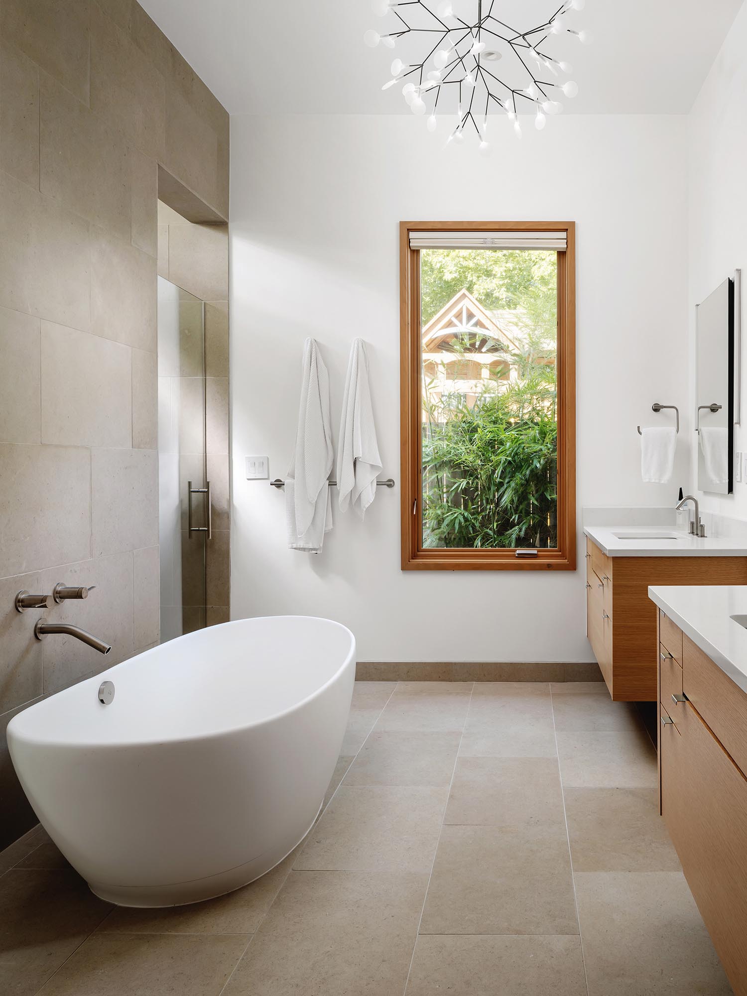 This en-suite bathroom includes a freestanding bathtub, a delicate chandelier, separate vanities, and limestone on the floor and wall.