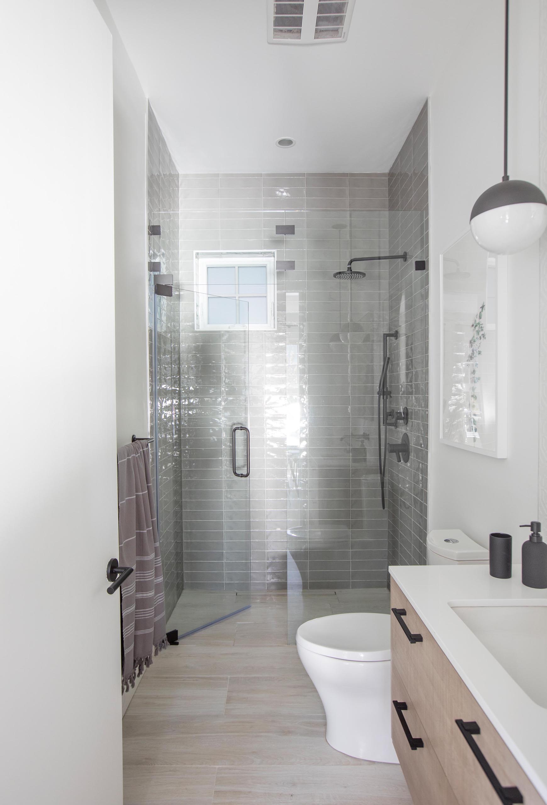 A modern bathroom that includes a wood vanity with a white countertop, and a shower lined with floor-to-ceiling gray tiles.