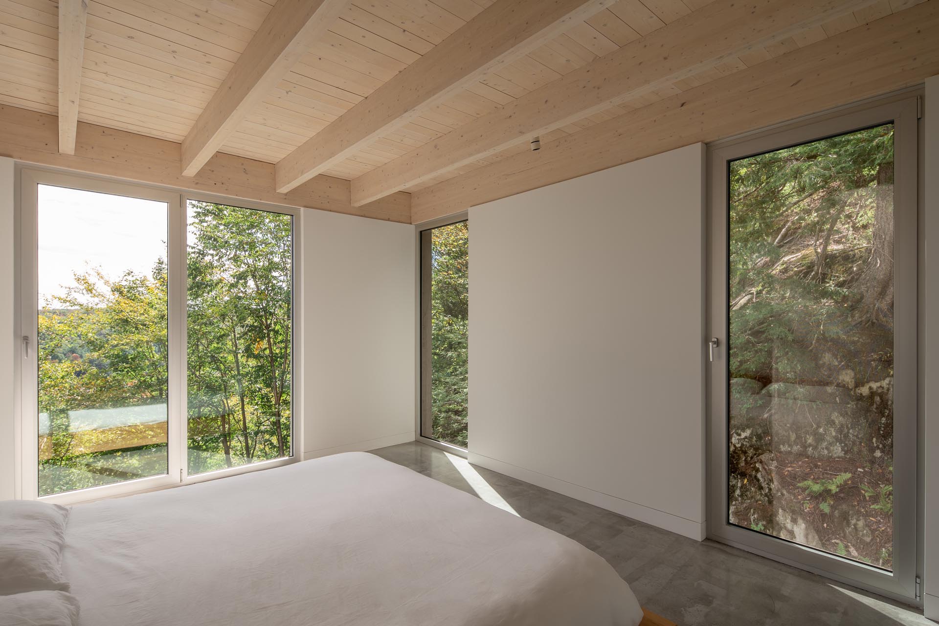 A master bedroom suite with minimal furniture has tall windows that frame the trees and rocks outside.