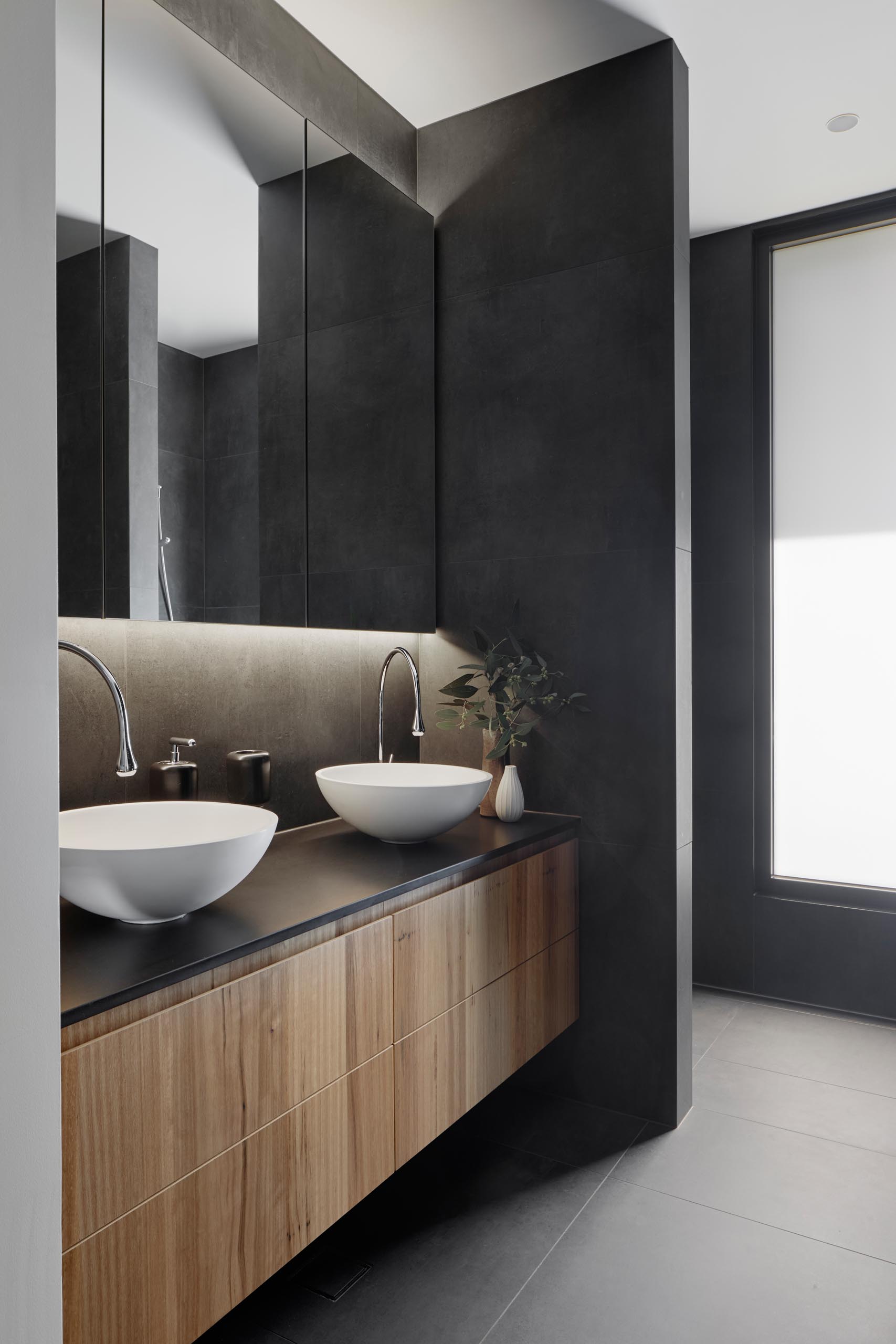 In this modern bathroom, dark gray walls and floors are combined with a wood vanity, white basins, and a backlit mirror.