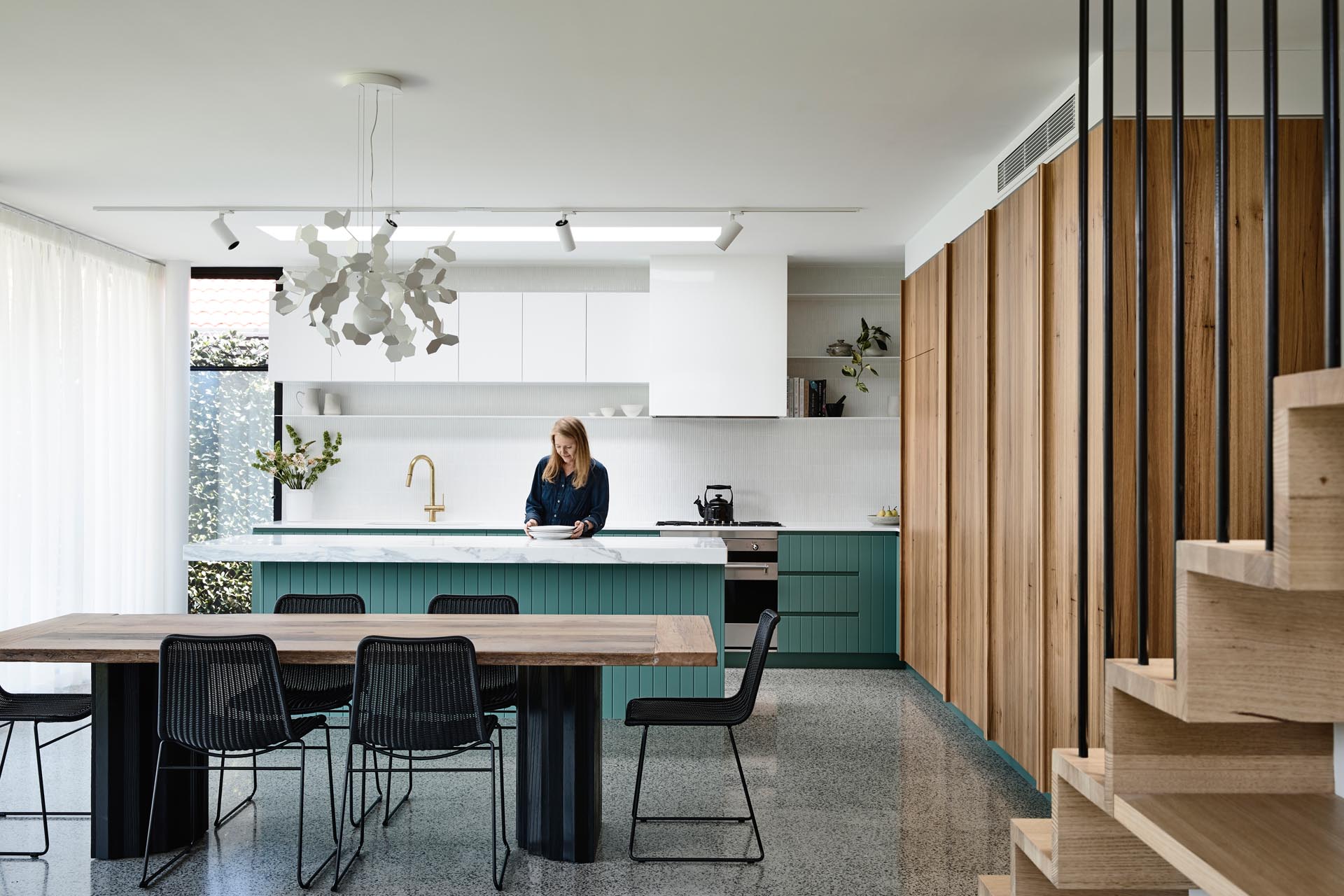 A modern interior with a dining area that has a large wood table with a black base that matches the black chairs, while in the kitchen, green cabinets add a colorful accent to the white countertops and upper cabinets.