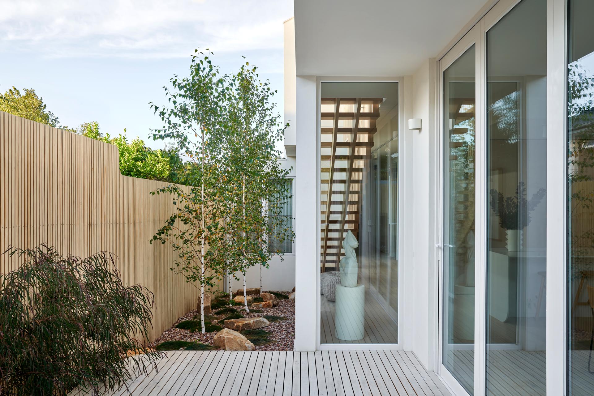 A modern house with a side garden that features a light wood fence, stones, grasses, and trees.