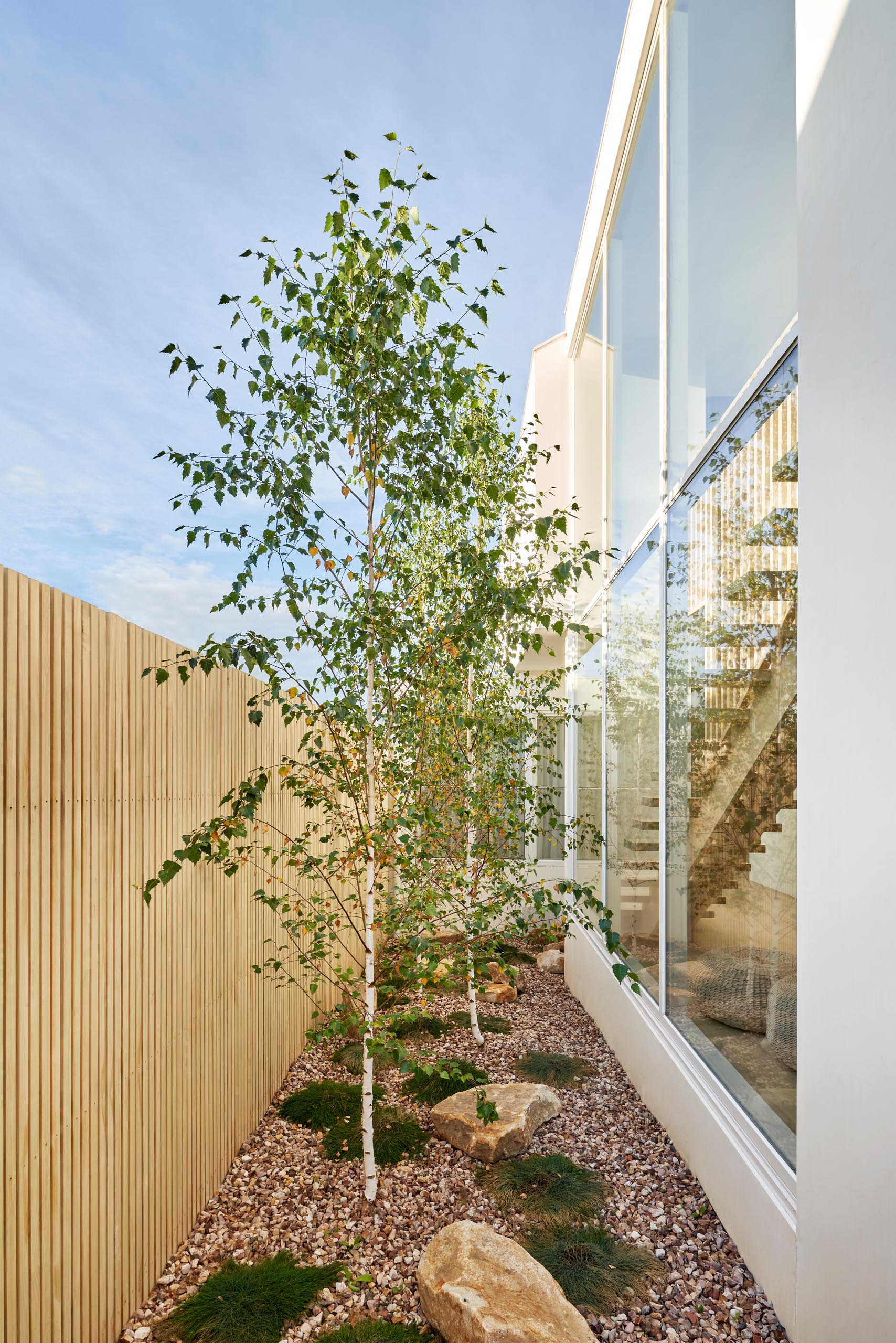 A modern house with a side garden that features a light wood fence, stones, grasses, and trees.