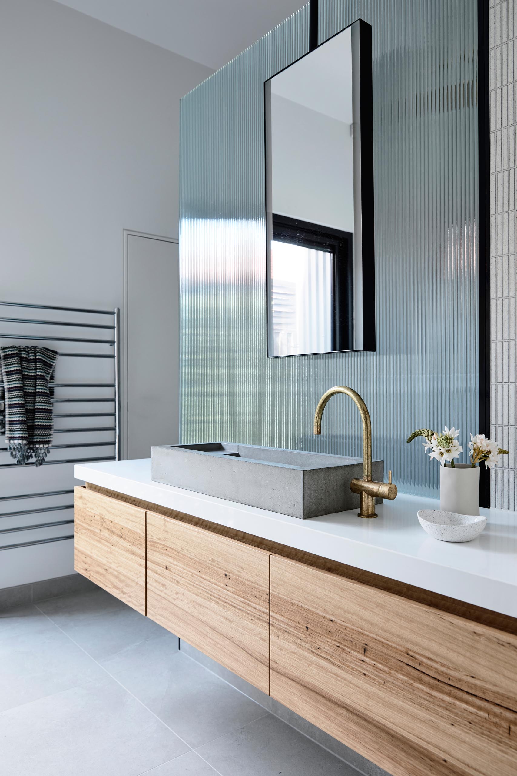 A modern bathroom with a long wood vanity, a concrete sink, bronze faucet, and a textured glass panel that hides the shower.