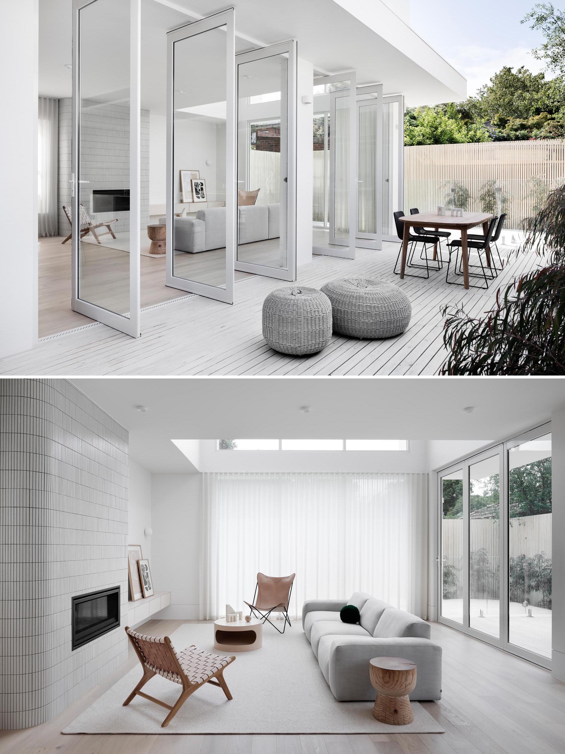 This modern house features pivoting glass doors, and and interior with a white tiled fireplace surround in the living room, a dining room with a light wood table, and a kitchen with minimalist cabinets and a white island.