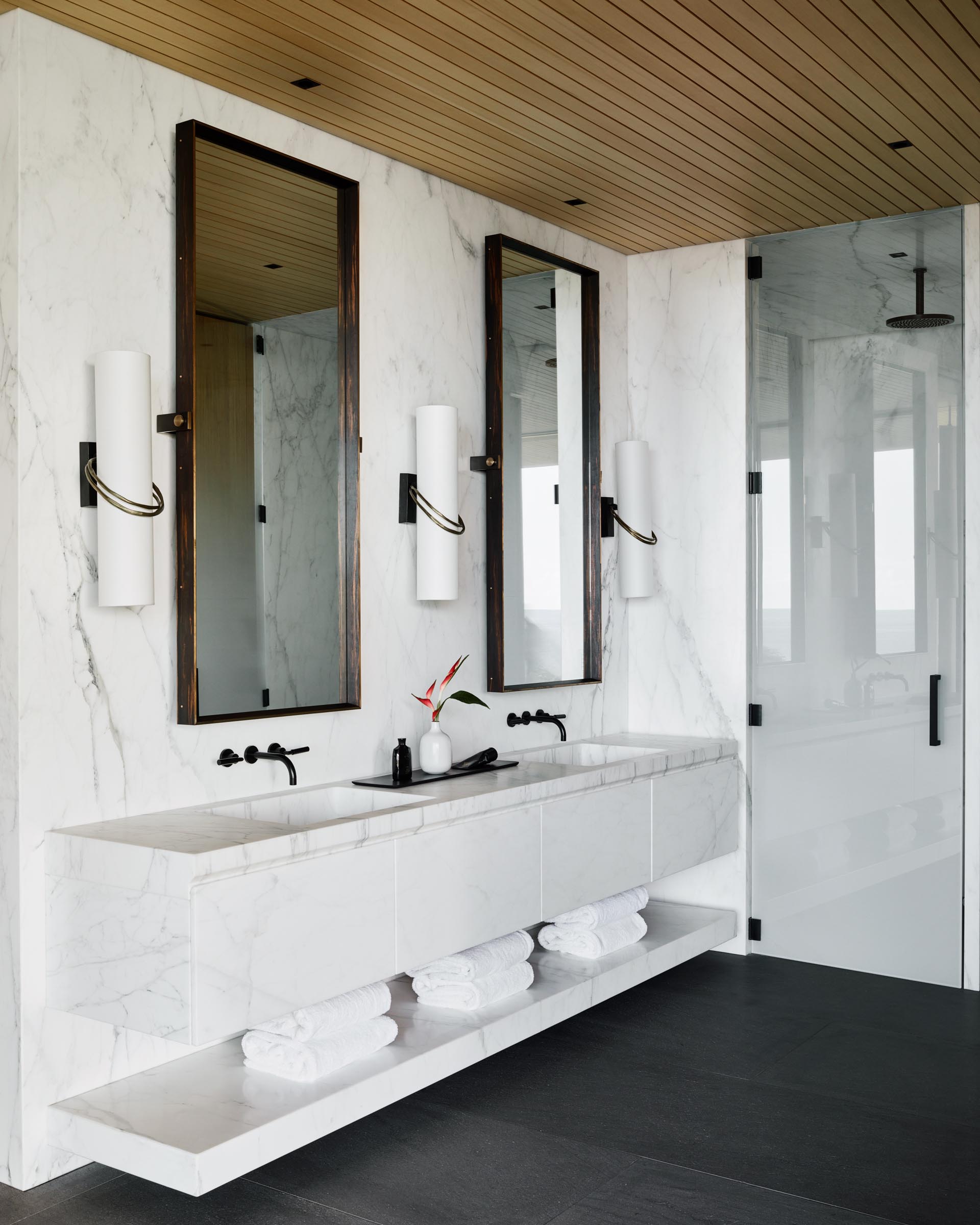 In the master bathroom, large format gray tiles cover the floor, vertical wood screens create privacy, and a white double-sink vanity floats above the ground and is adjacent to the glass-enclosed shower.