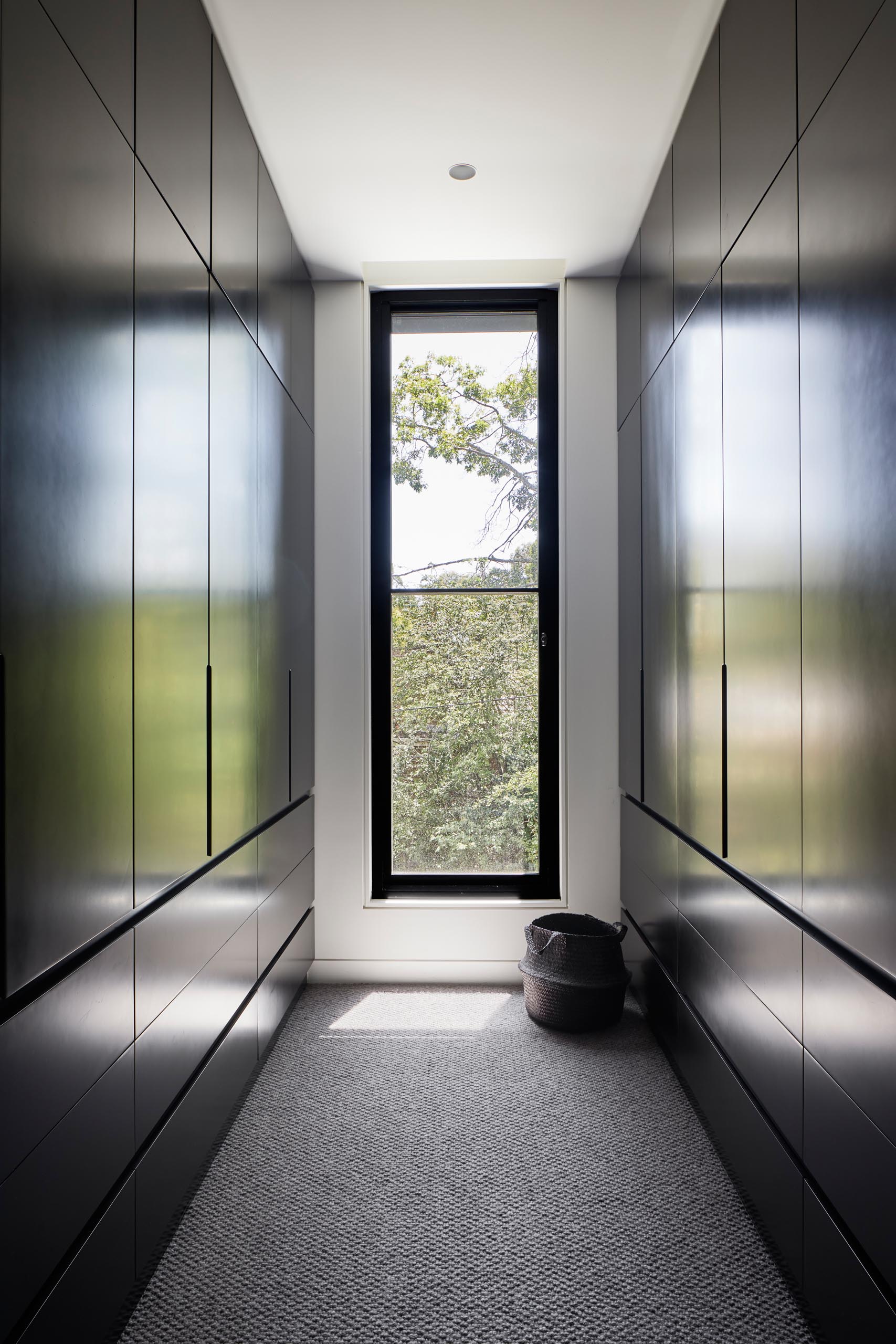 In this modern walk-in closet, there's dark floor-to-ceiling cabinetry that lines the walls, and allows the eye to focus on the tree view through the window.