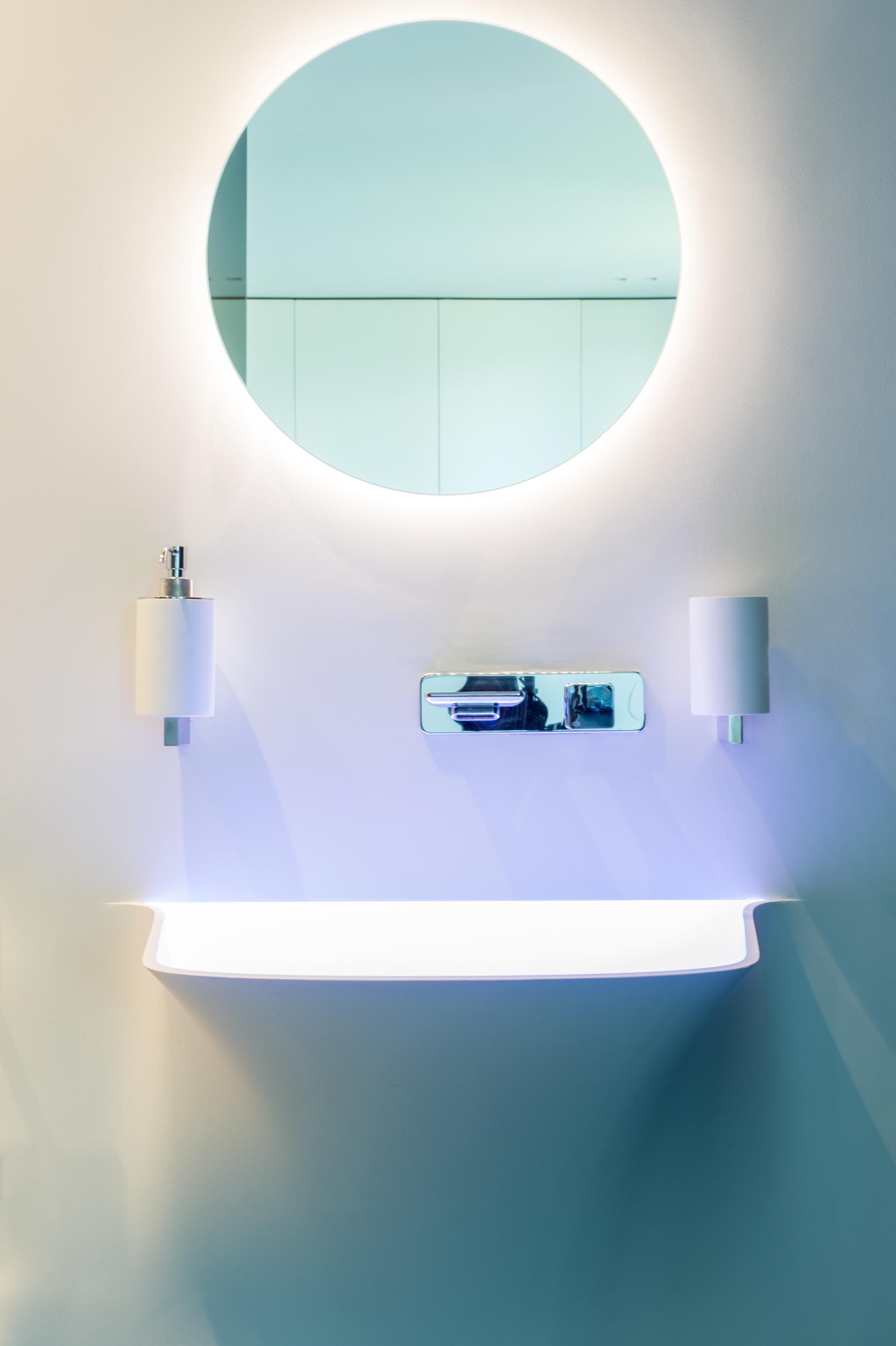 A modern bathroom with a seamless white vanity and backlit round mirror.