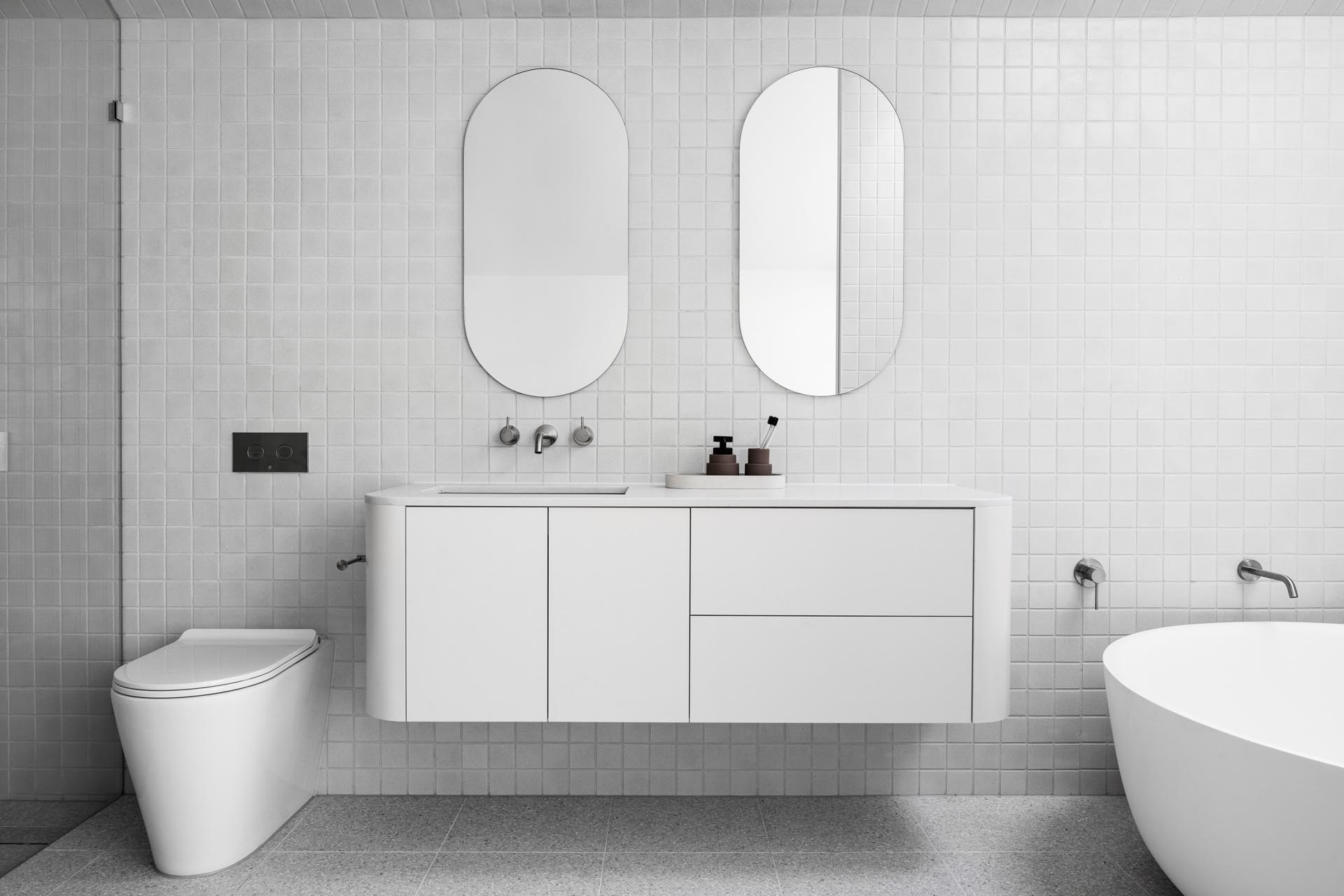 In this modern bathroom, square gray tiles cover the walls and floor, while pill-shaped mirrors hang above the floating white vanity with curved corners. One end of the bathroom is dedicated to the shower, while the other has a round freestanding bathtub located beneath a skylight.