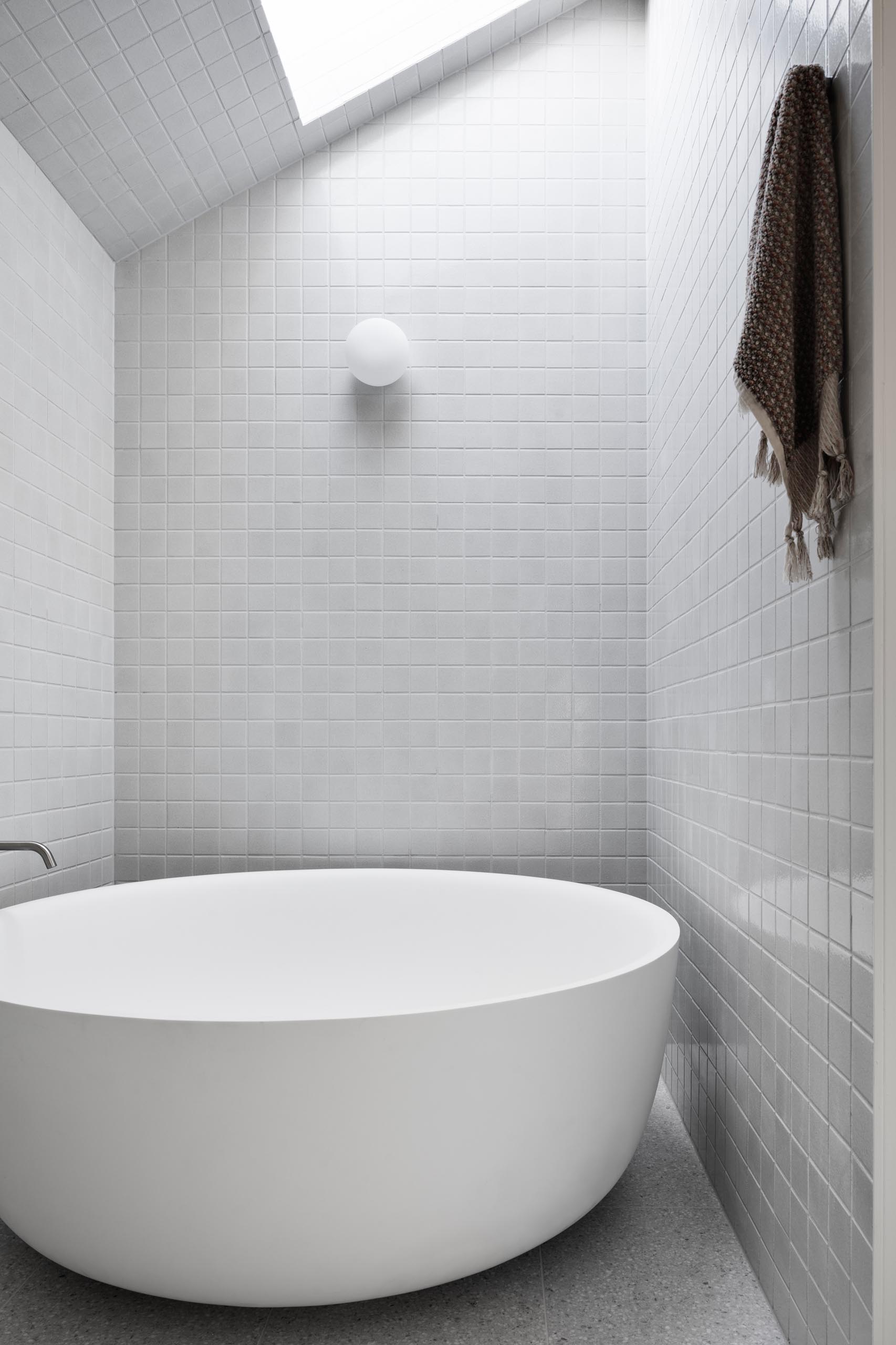 In this modern bathroom, square gray tiles cover the walls and floor, while pill-shaped mirrors hang above the floating white vanity with curved corners. One end of the bathroom is dedicated to the shower, while the other has a round freestanding bathtub located beneath a skylight.