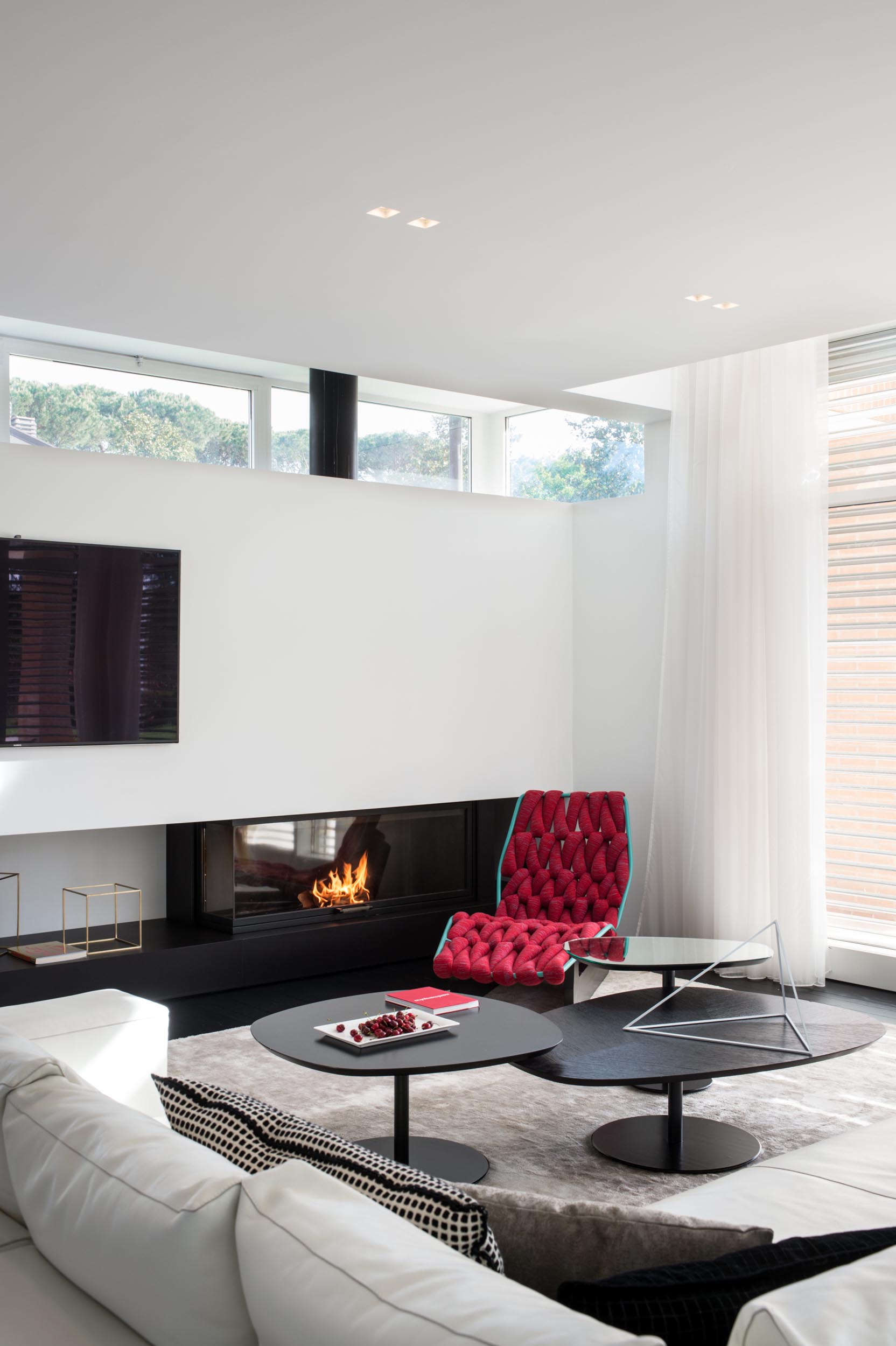 In this modern living room, a white corner sofa sits on top of a large rug, while black coffee tables complement the black fireplace. A woven red chaise has been chosen to add a touch of color.