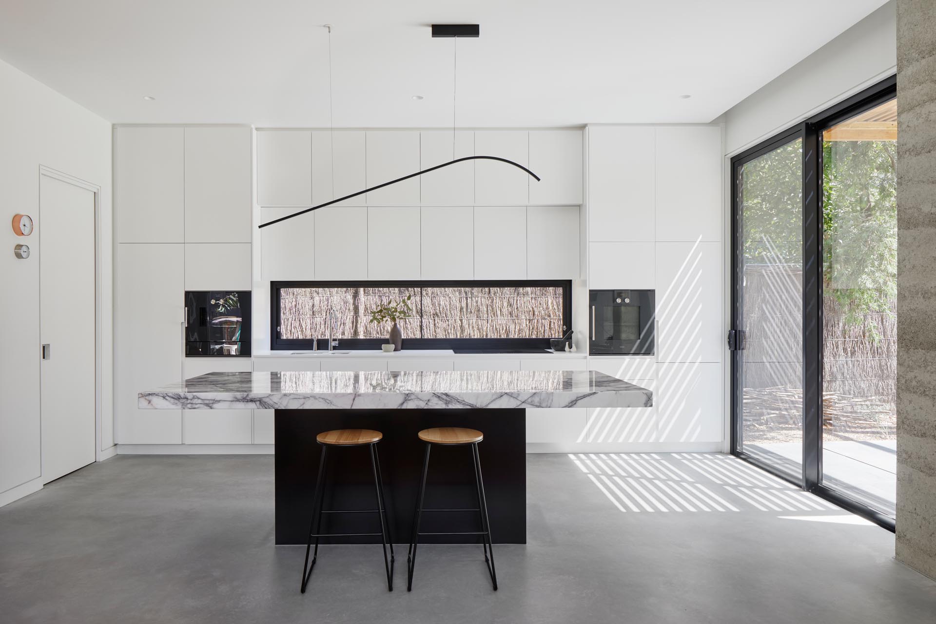 In this modern kitchen, minimalist white cabinets with an integrated fridge and freezer, contrast the built-in appliances, horizontal black window frame, and the black island base. The island has a thick marble countertop, while pure white countertops are featured along the wall.