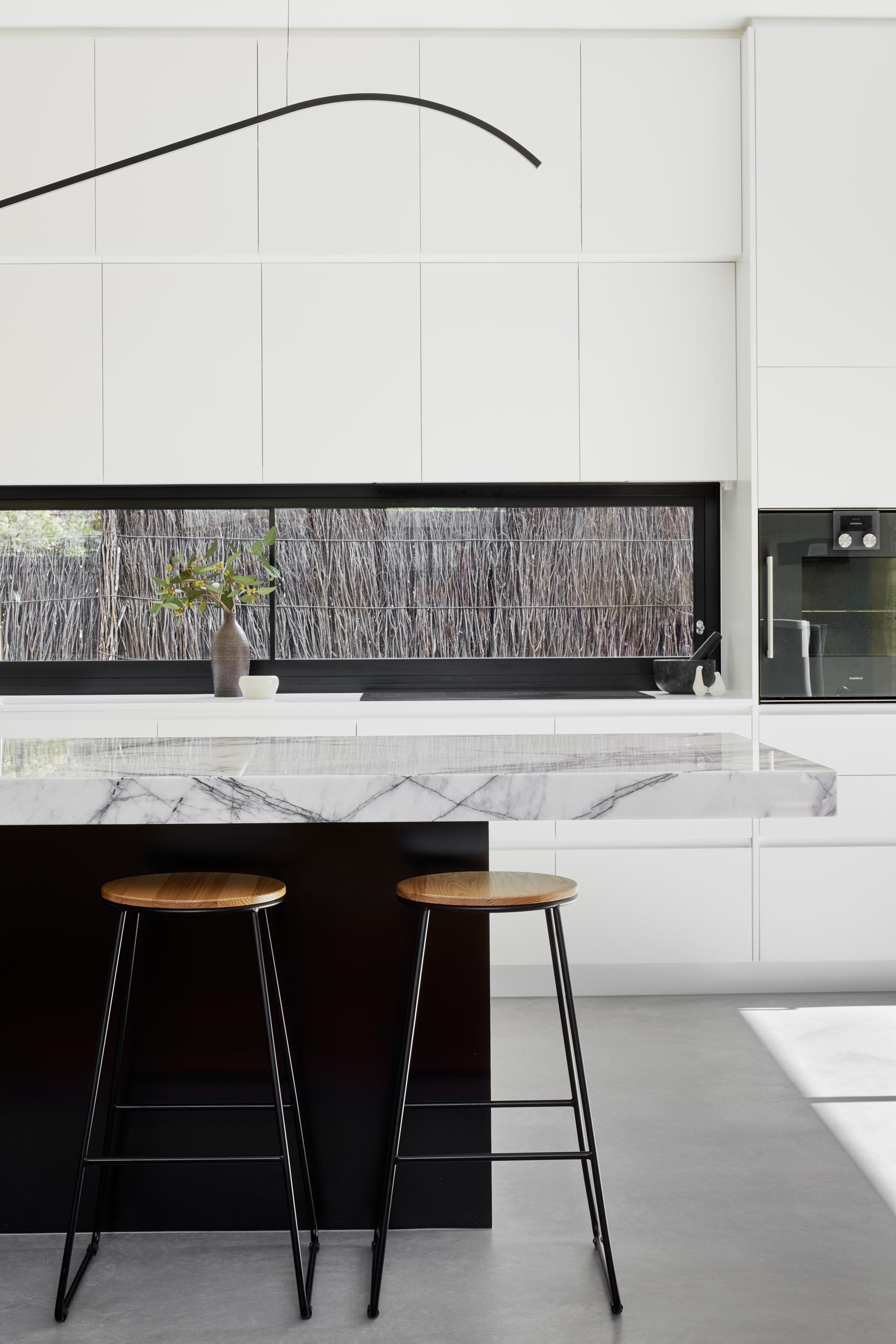 In this modern kitchen, minimalist white cabinets with an integrated fridge and freezer, contrast the built-in appliances, horizontal black window frame, and the black island base. The island has a thick marble countertop, while pure white countertops are featured along the wall.