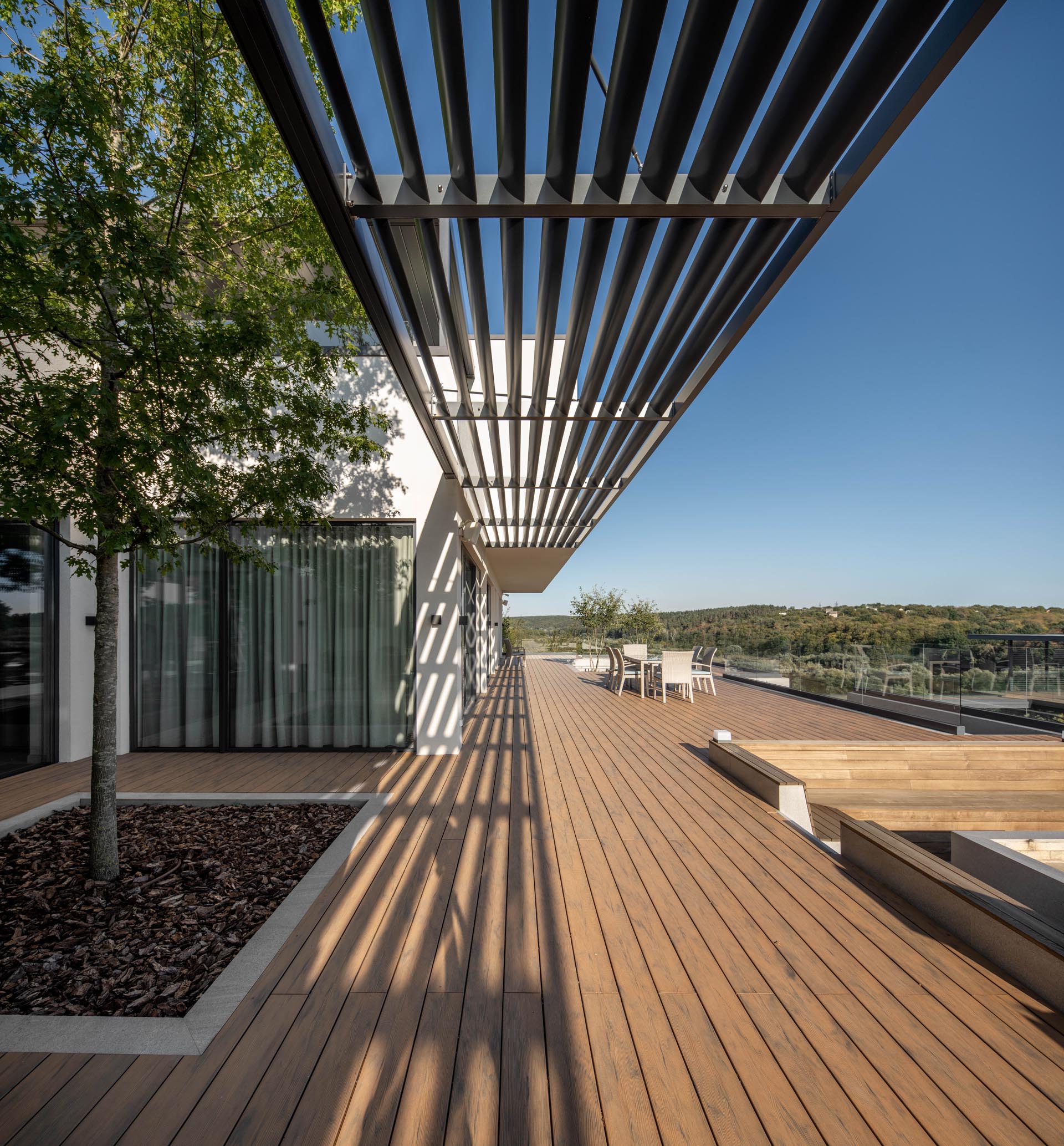 A modern house with a roomy terrace that's designed as a relaxing zone with lounges, a dining zone, and a small garden with trees, bushes and lawn.