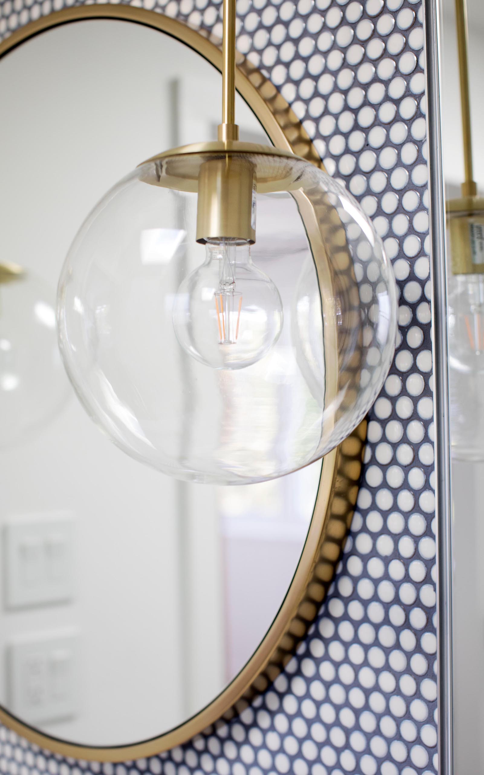 A modern bathroom with white penny tiles, contrasting dark grout, a round mirror with gold frame, and matching glass ball pendant light.