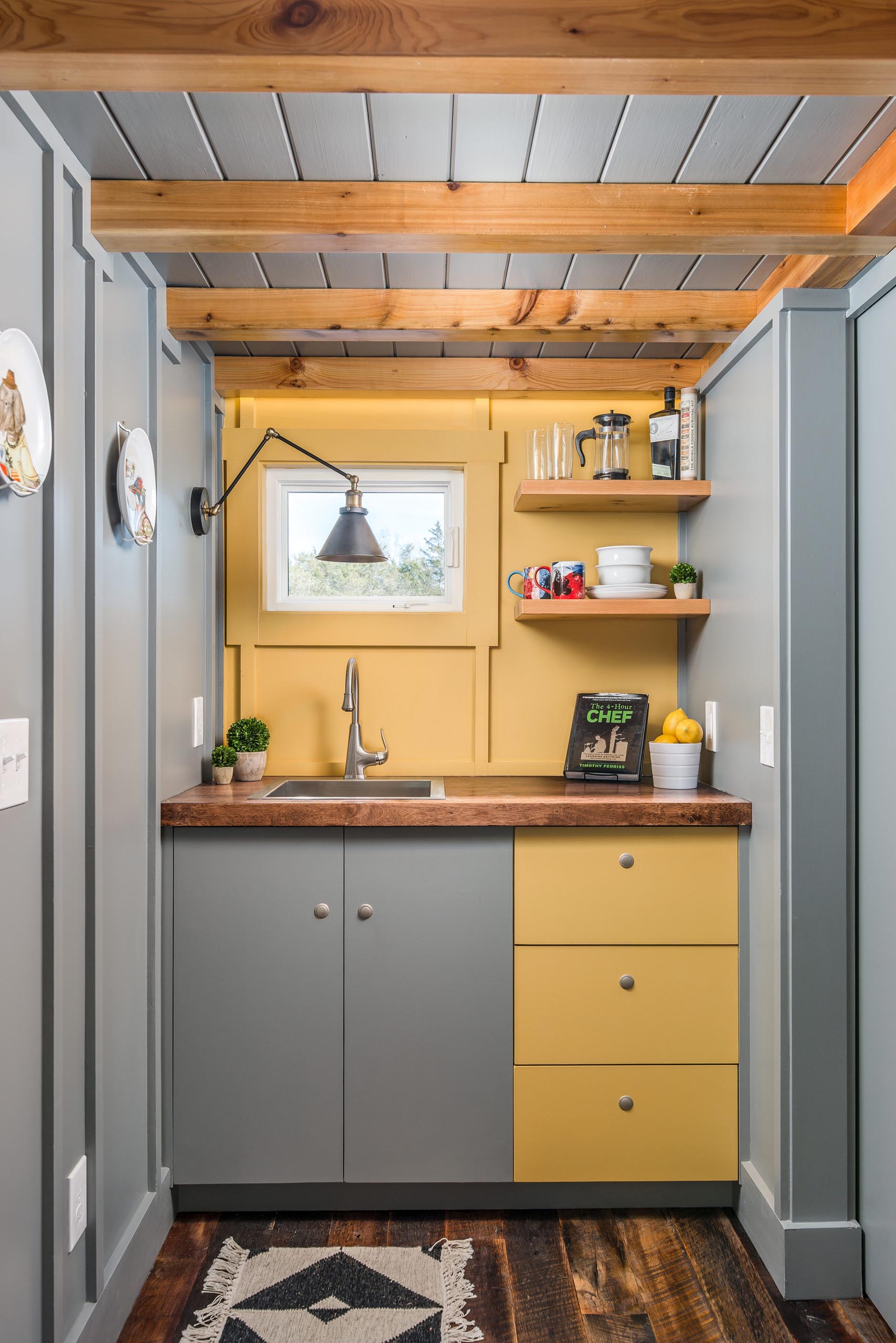 The soft yellow, gray, and wood kitchen of this tiny home includes a stainless steel faucet and matching sink, a dark wood countertop, and floating corner shelves.