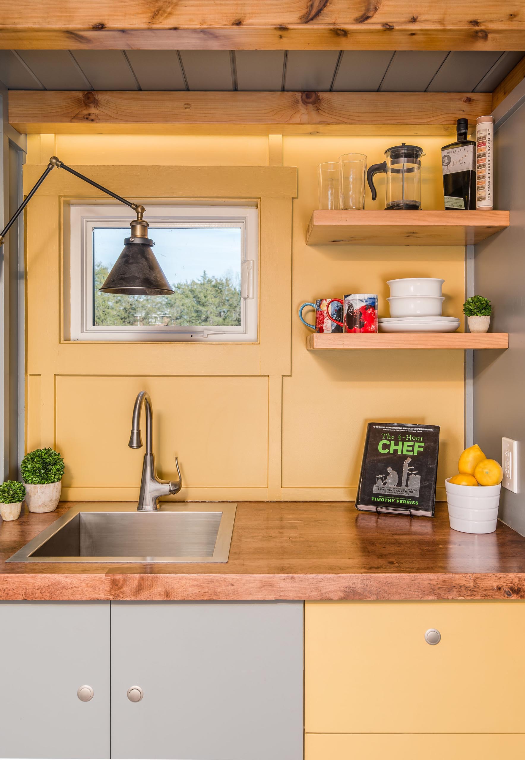 The soft yellow, gray, and wood kitchen of this tiny home includes a stainless steel faucet and matching sink, a dark wood countertop, and floating corner shelves.