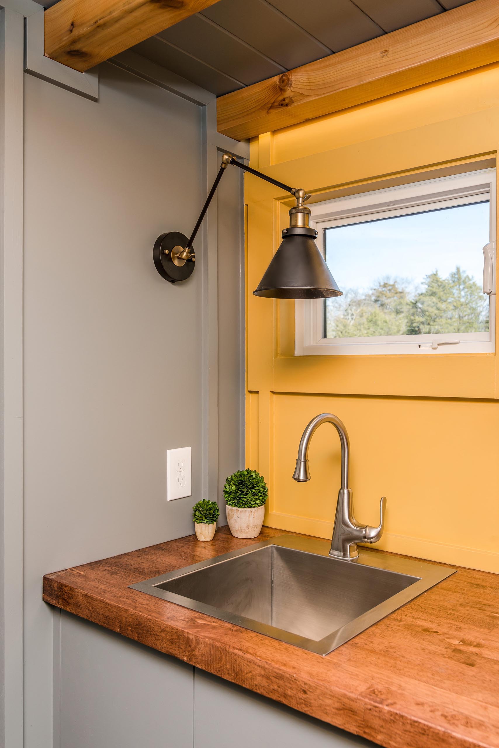 The soft yellow, gray, and wood kitchen of this tiny home includes a stainless steel faucet and matching sink, a dark wood countertop, and floating corner shelves.