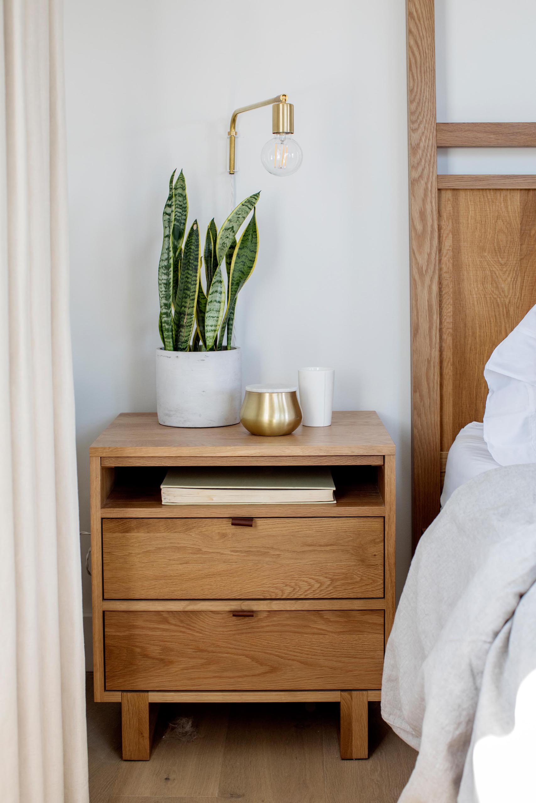 A contemporary wood bedside table with a plant and wall lamp.