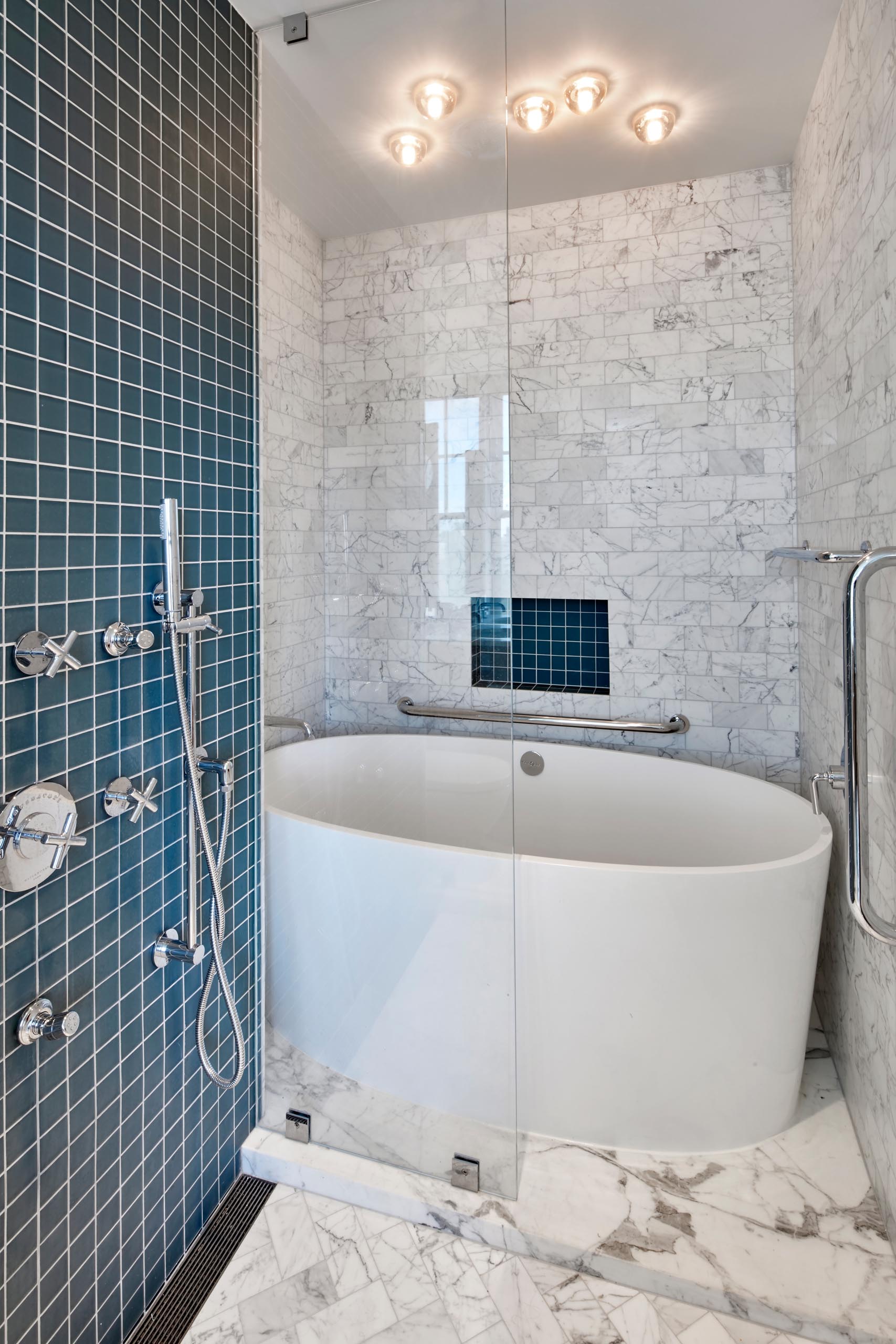 A modern bathroom with white soaking tub, gray marble subway tiles, and shower and a shelving niche lined with square turquoise tiles.