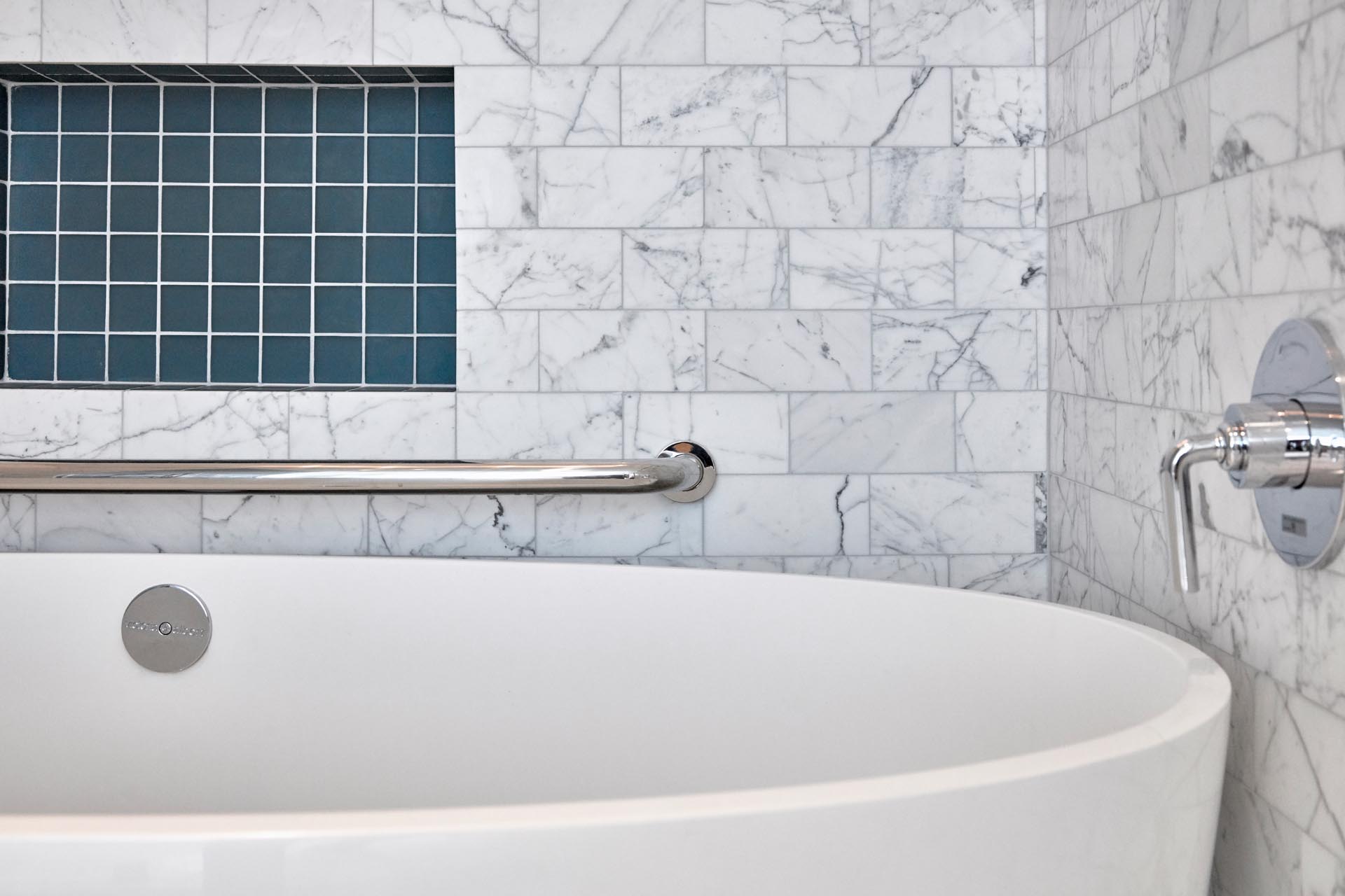 A modern bathroom with white soaking tub, gray marble subway tiles, and a shelving niche lined with square turquoise tiles.