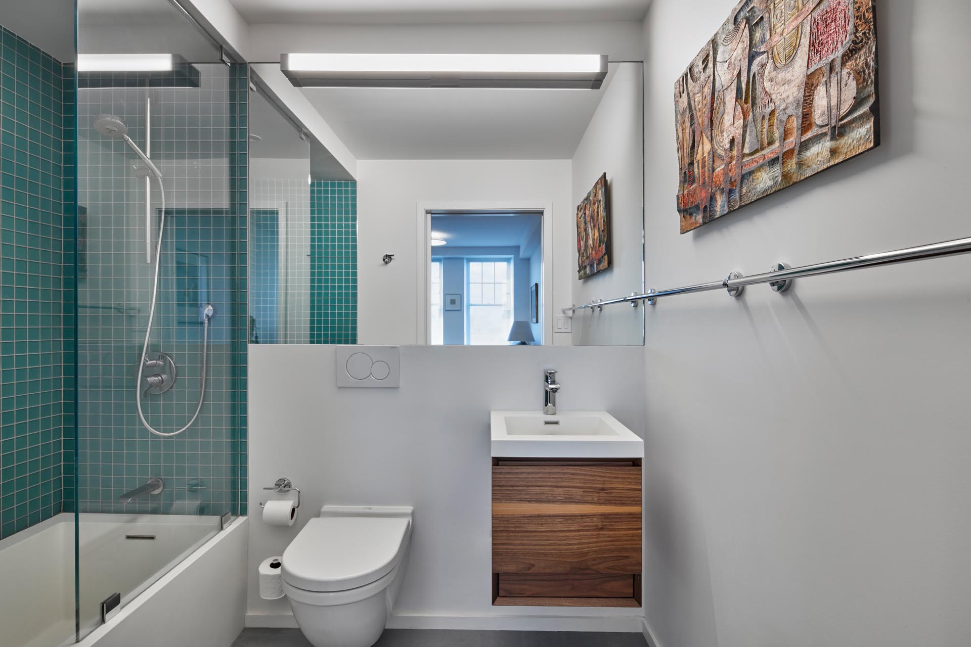 A modern bathroom with bright white walls, a simple wood vanity with white sink, and a shower/bath combo with square turquoise tiles on the walls.