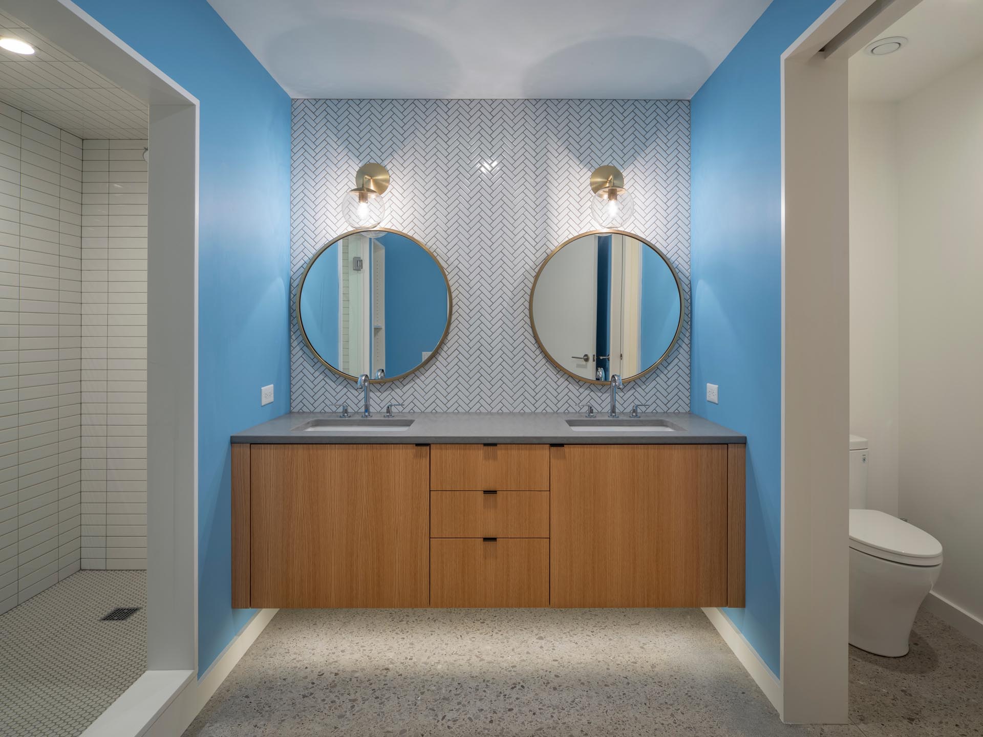 A modern bathroom with bright blue walls that add a pop of color, white a floating wood vanity has lighting underneath it, and above, there are two round mirrors with bronze frames that match the sconces.