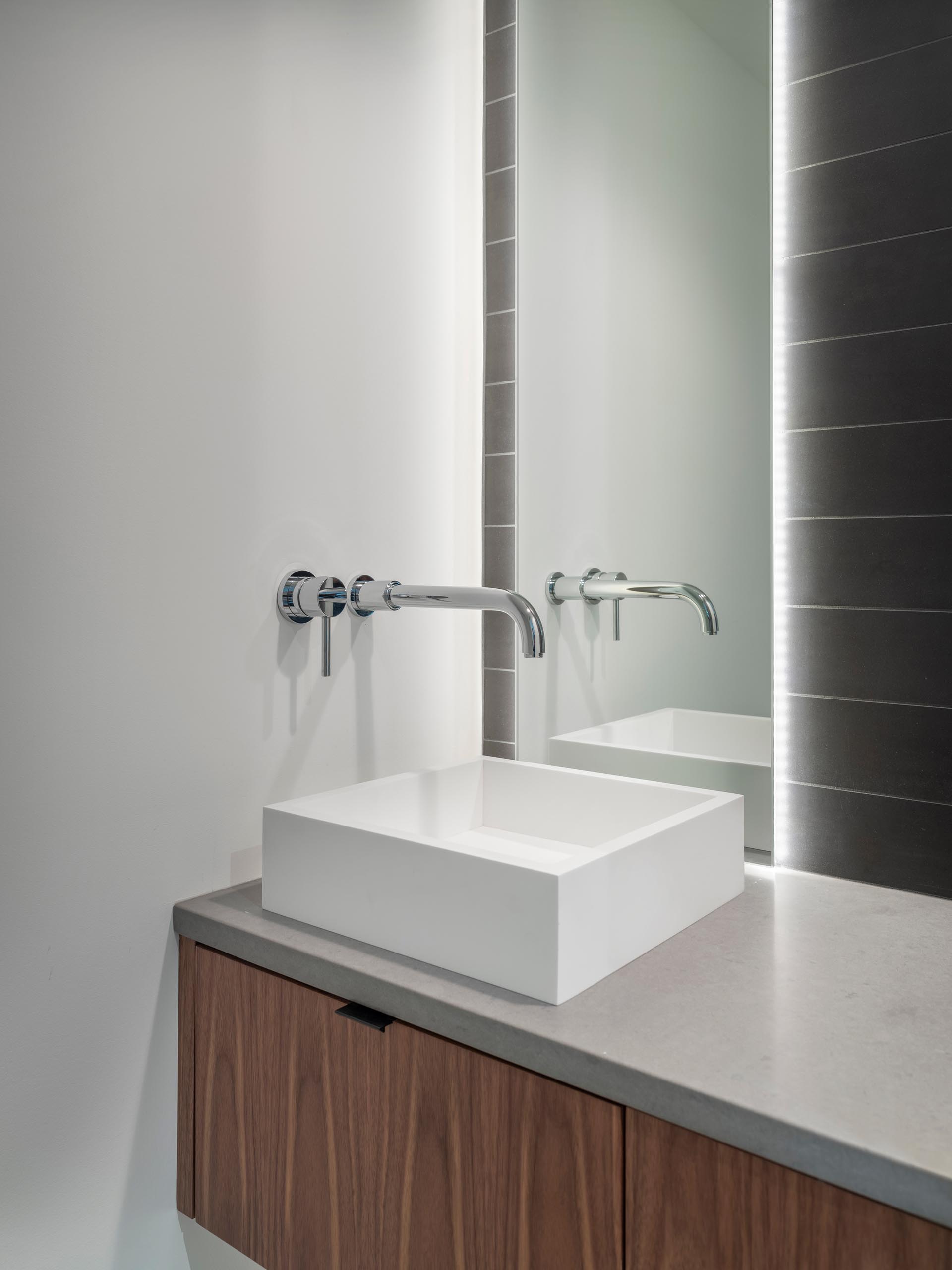 A modern bathroom with a dark tile wall that's lit up by the LED lighting that's behind the mirror.