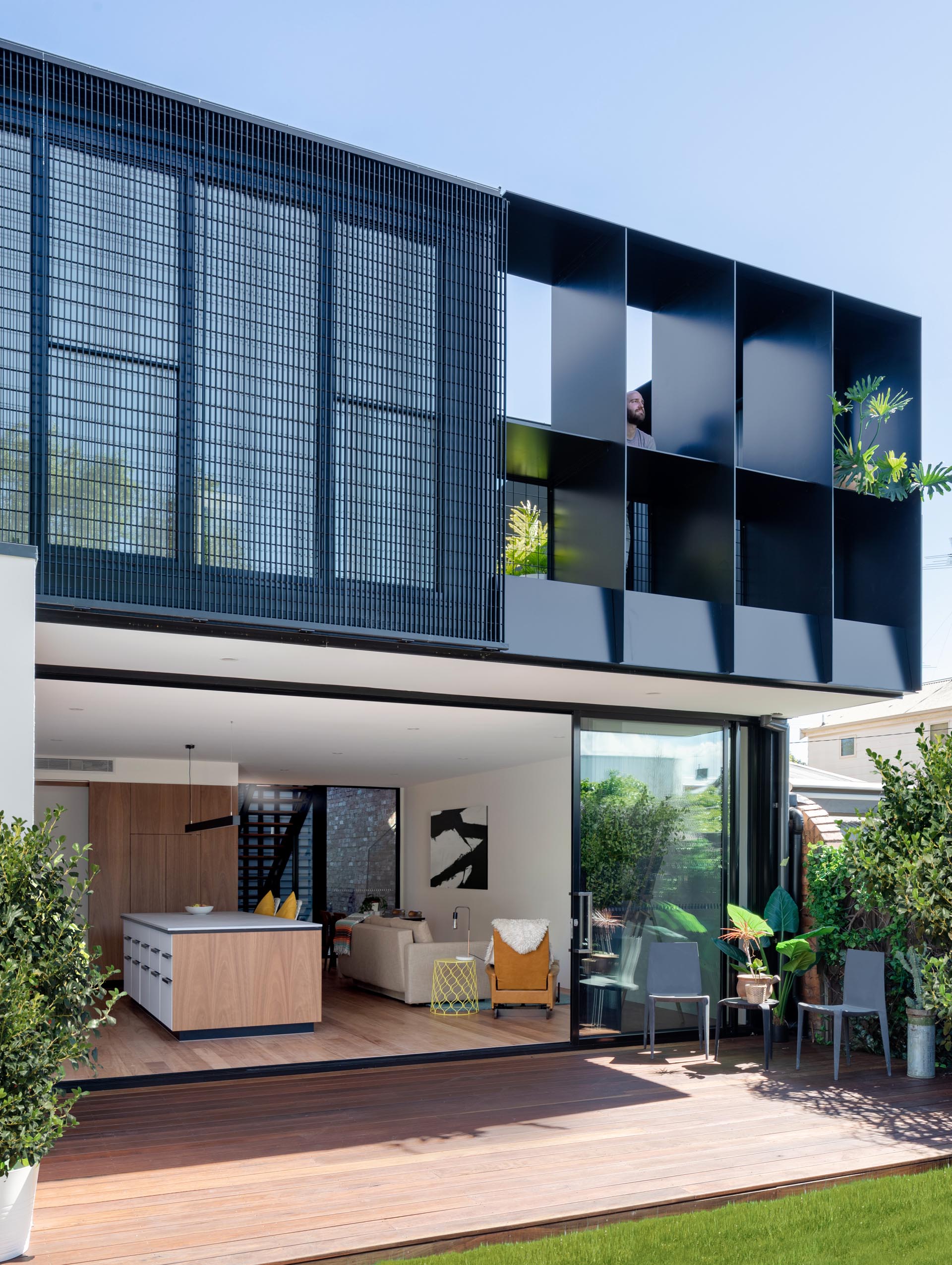 A modern house extension with a sliding glass wall that opens to the backyard, which provides a view of the rear dark gray corrugated metal extension.