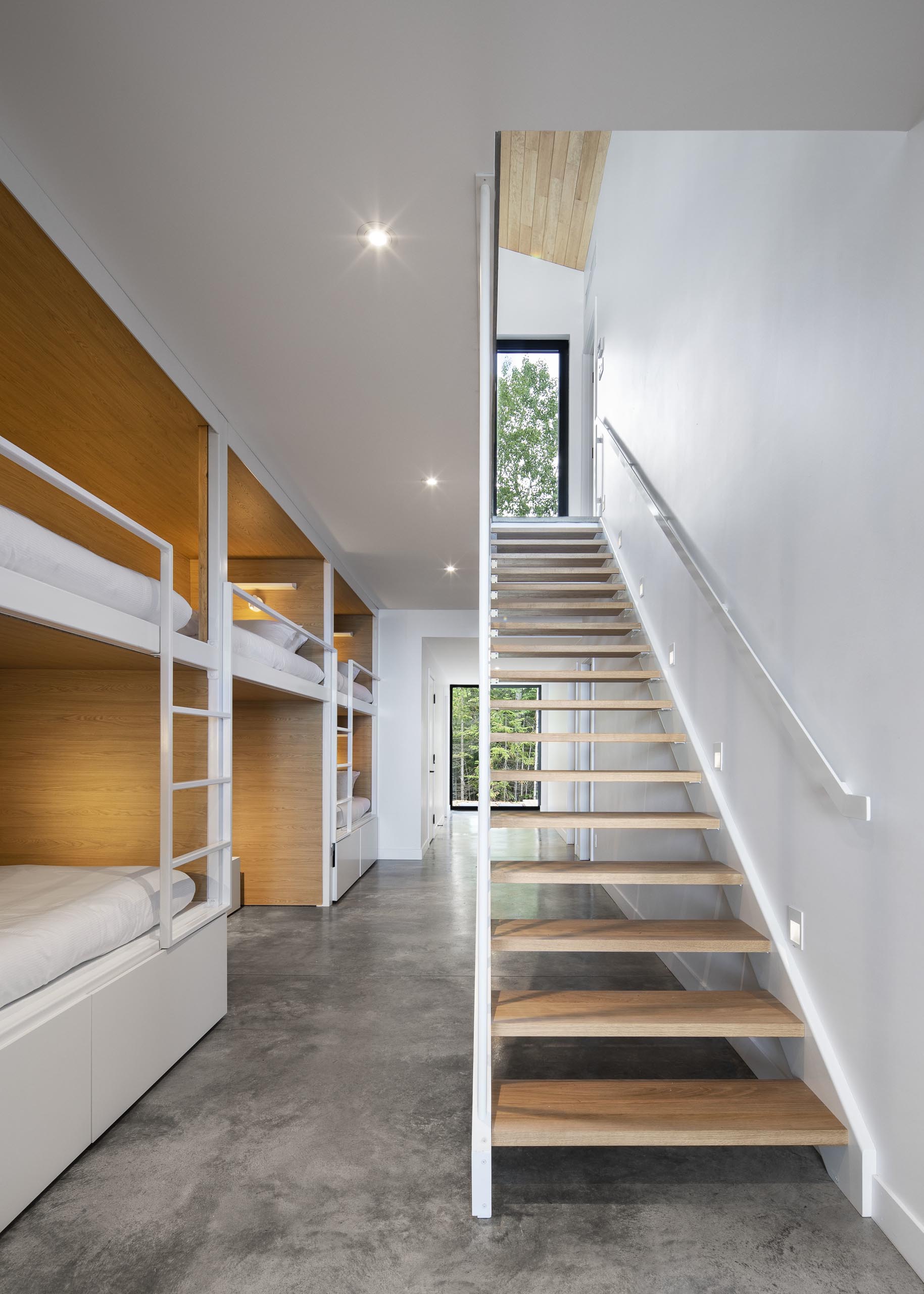 At the bottom of these modern stairs stairs and open to the hallway, are multiple sets of bunk beds. These bunk beds have a wood interior, with white suppers and ladders that match the walls.