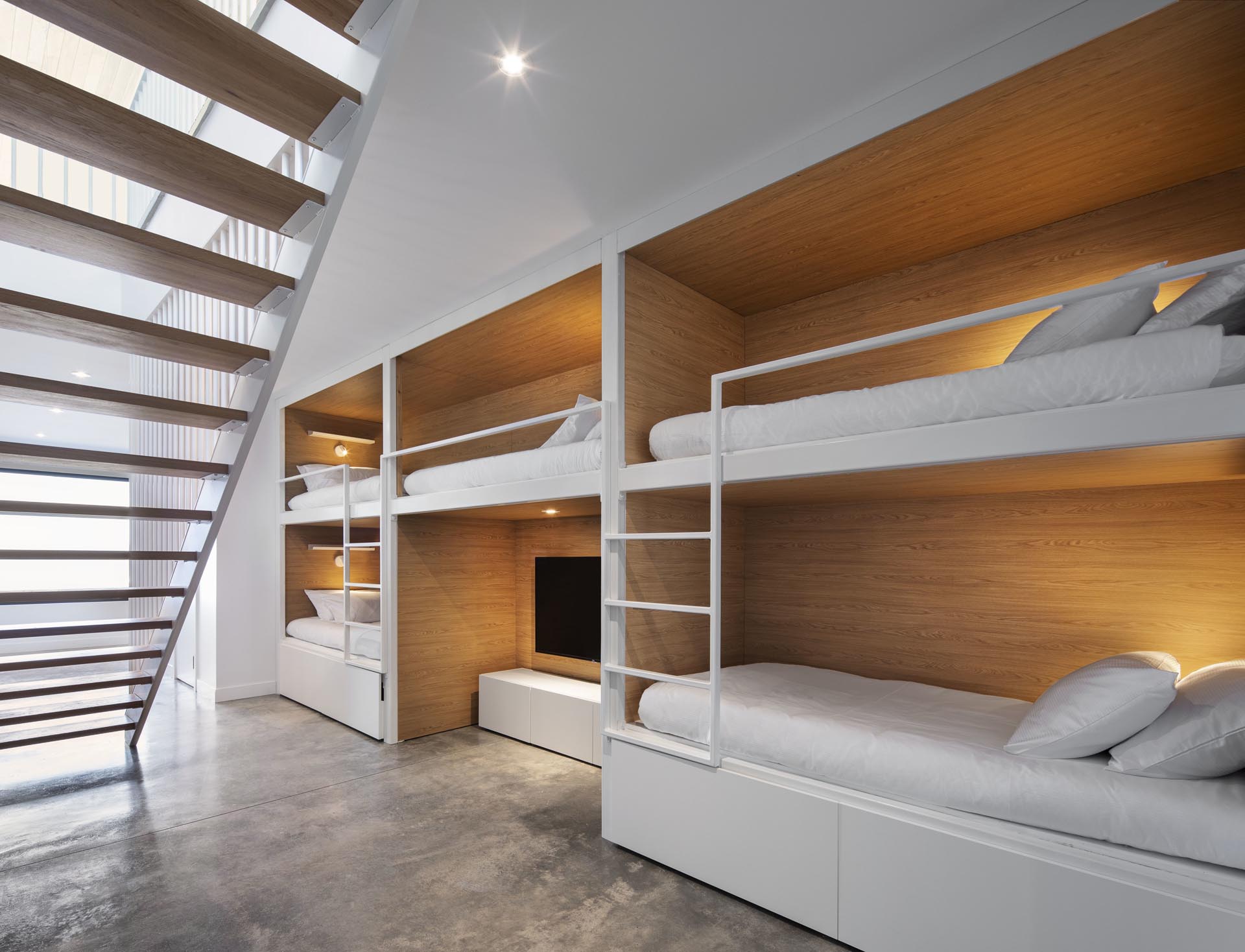 At the bottom of these modern stairs stairs and open to the hallway, are multiple sets of bunk beds. These bunk beds have a wood interior, with white suppers and ladders that match the walls.