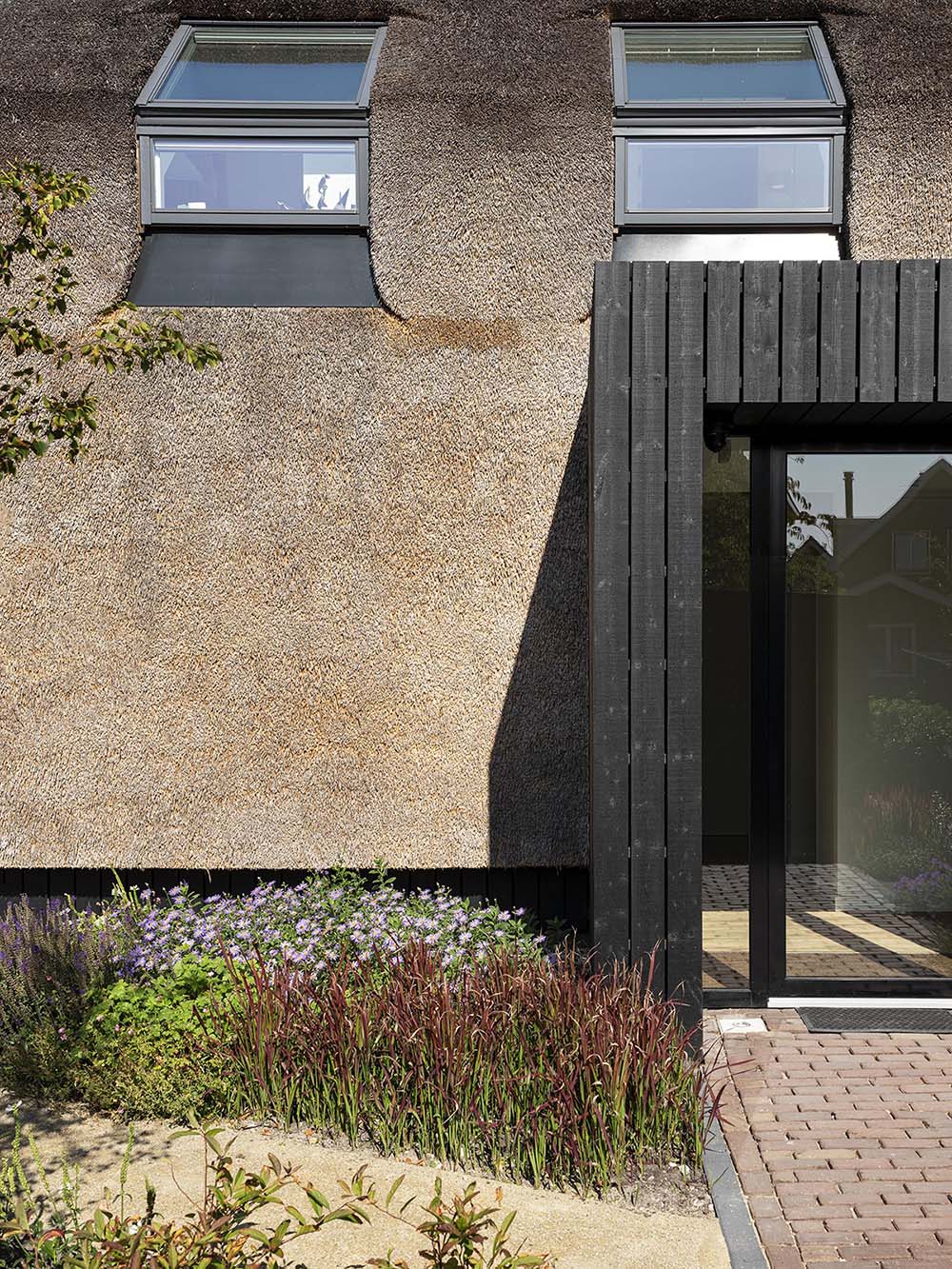 The exterior of this modern house is covered in thatch, which runs vertically down from the rooftop and along the sides of the house. The thatched roof is contrasted by wood with a black finish.