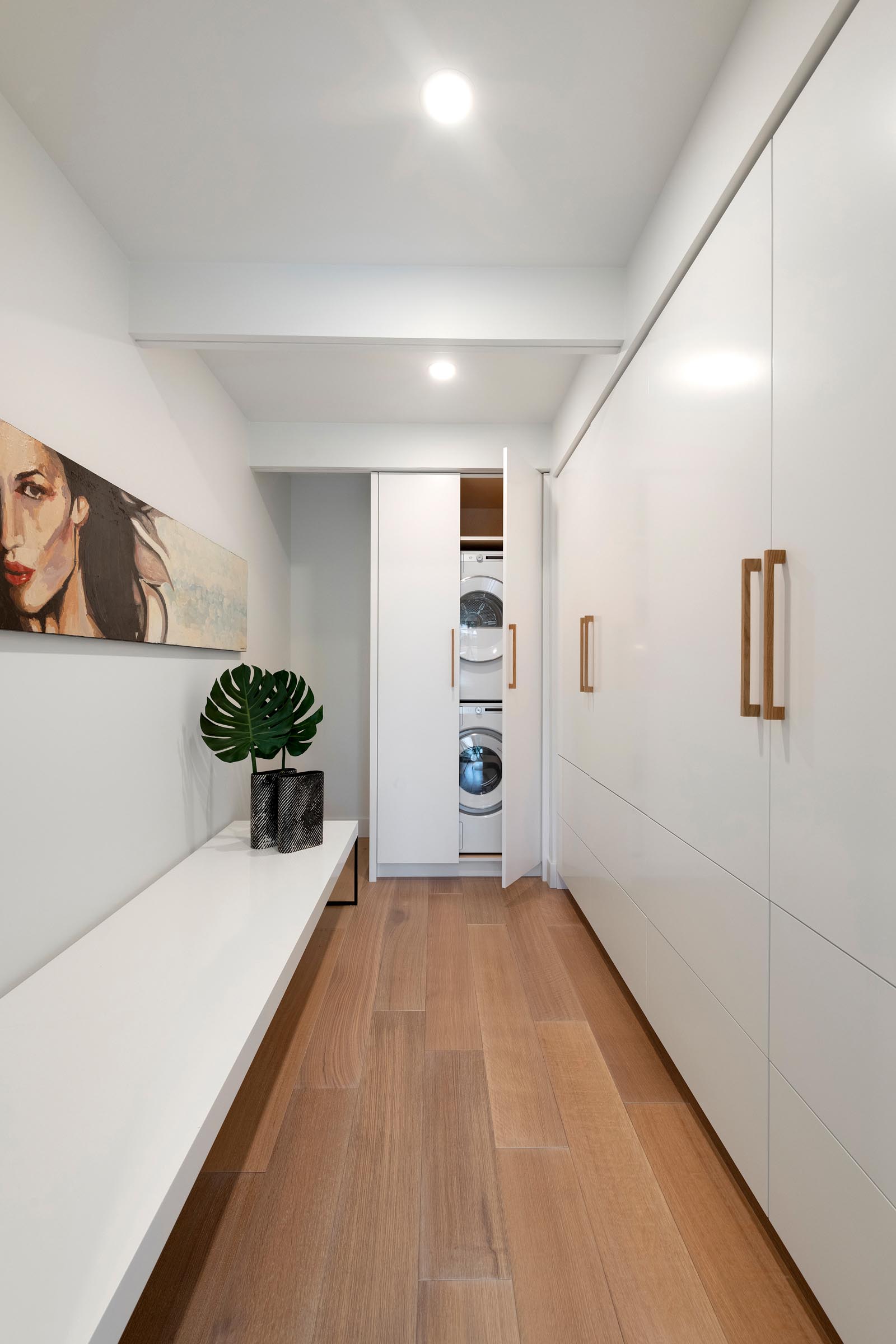 In this modern white laundry room, there's a long bench and wall of cabinetry with a washer and dryer hidden in the end cabinet.