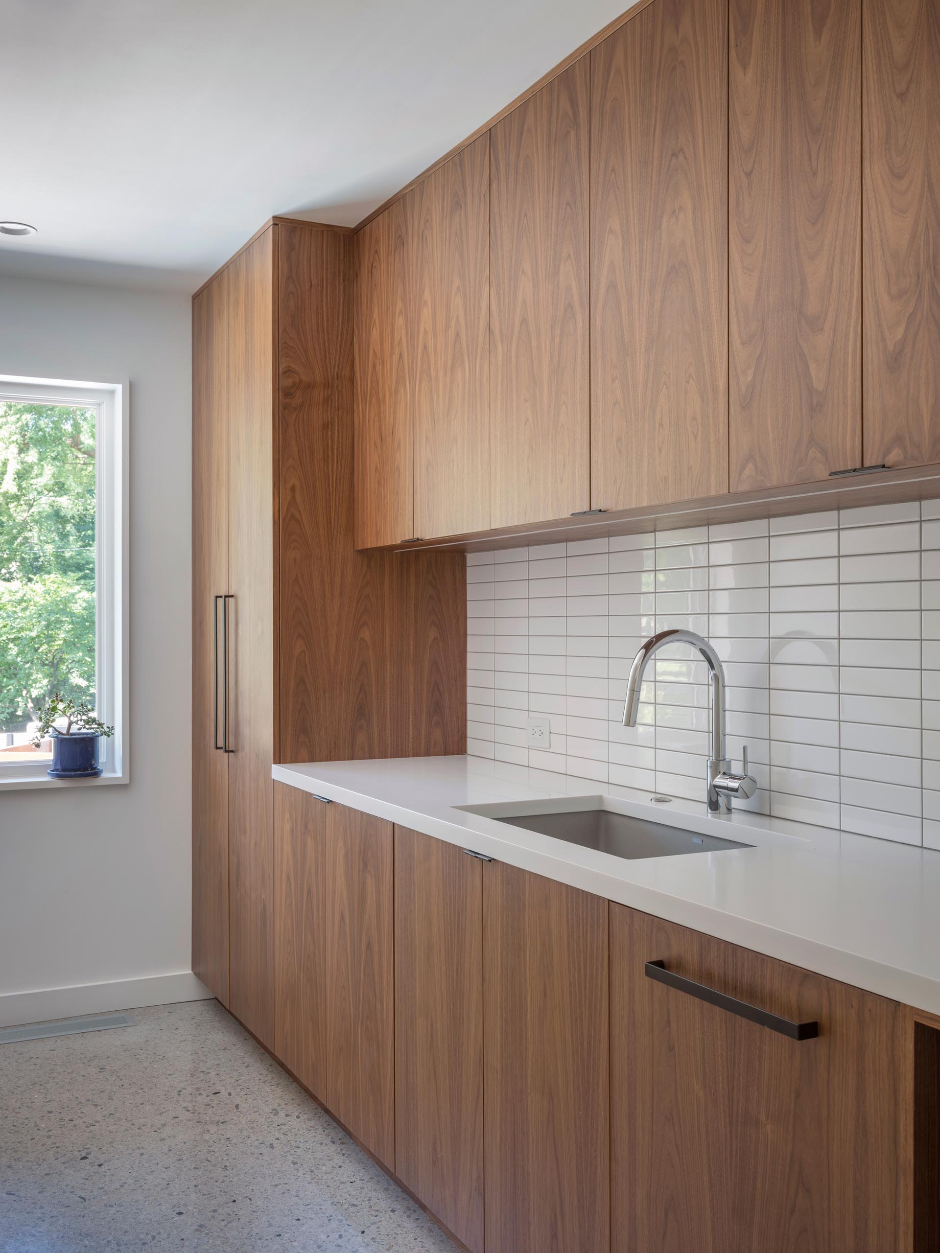 A modern laundry room with wood cabinets, a white countertop, undermount sink, and white subway tiles.