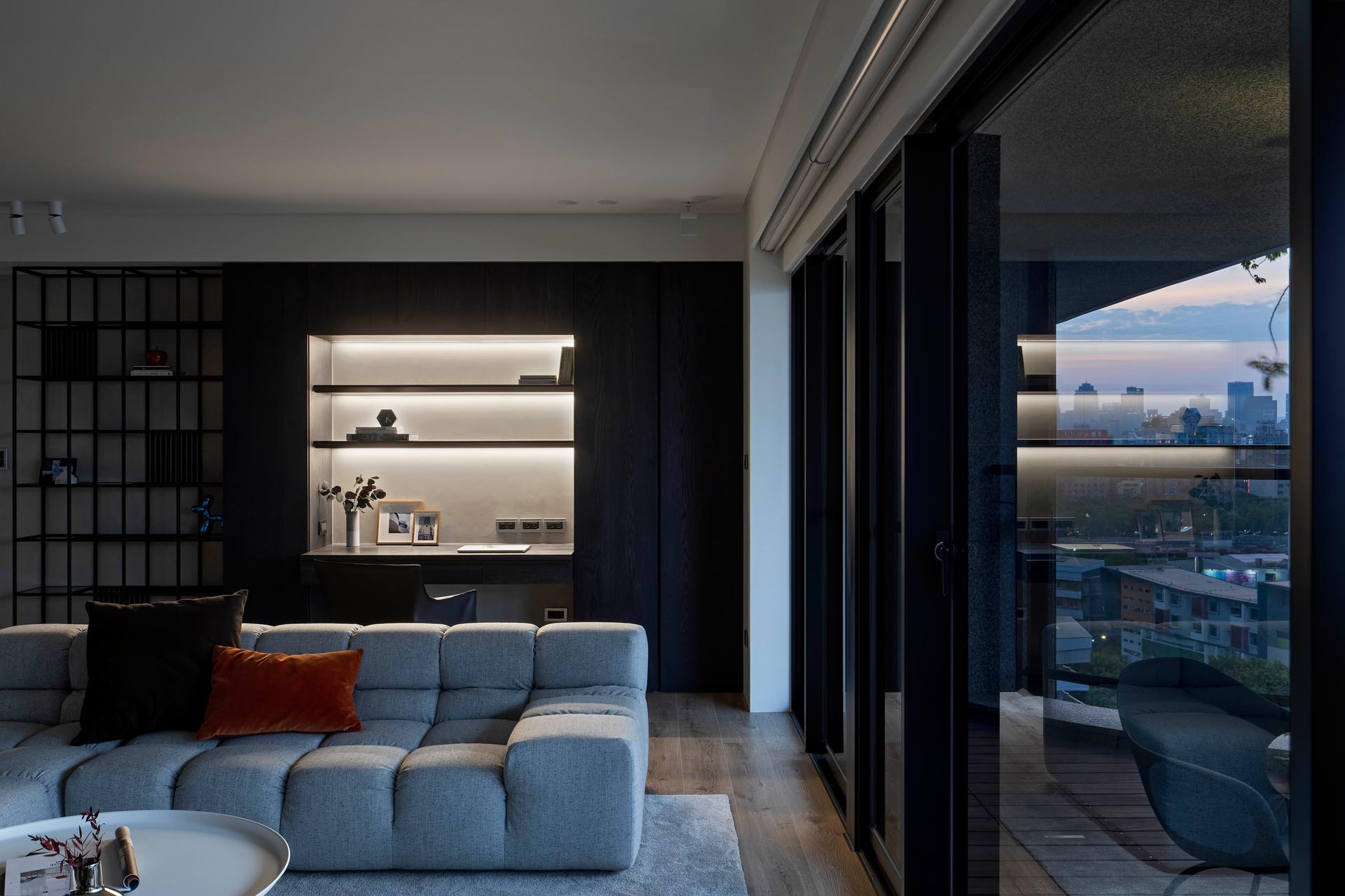 A modern living room with a wall dedicated to a home office, that includes built-in shelving that surrounds a desk and hidden LED lighting.