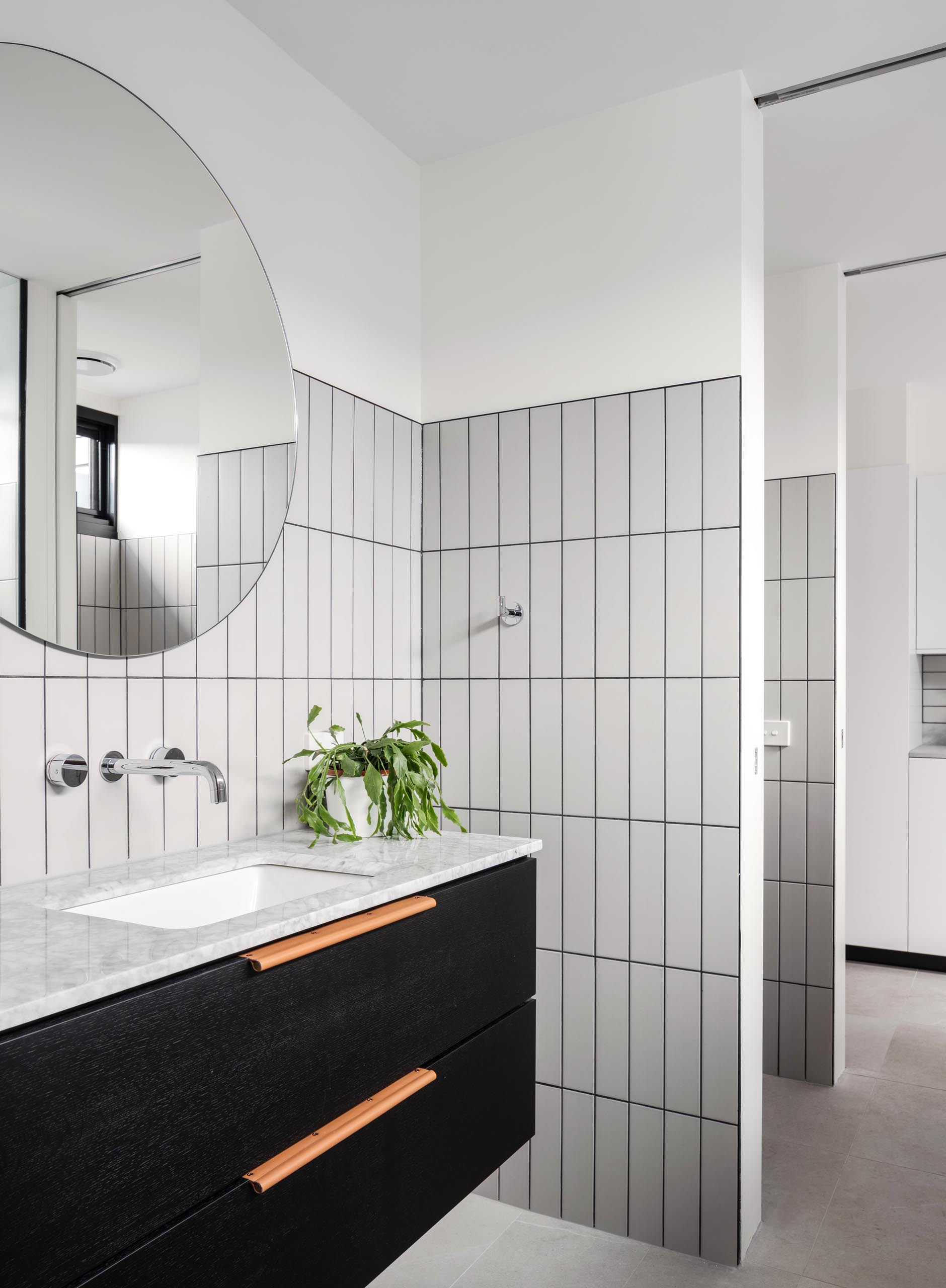 In the bathroom, rectangular light gray tiles with dark grout have been laid in a vertical pattern, while a black vanity sits below a round mirror.