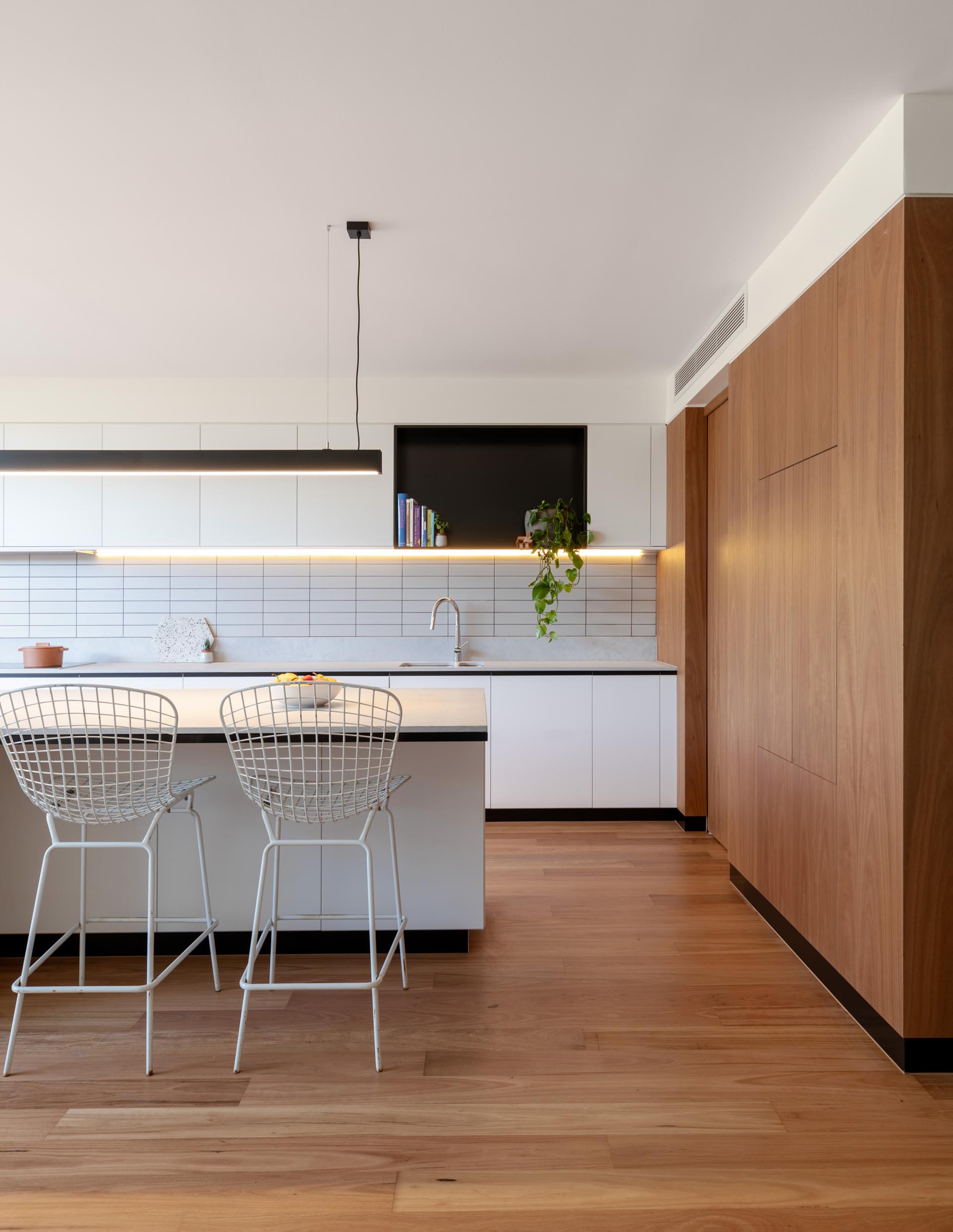 A modern kitchen includes minimalist white cabinets, a black accent shelf, a large island with room for seating, and undermount lighting.