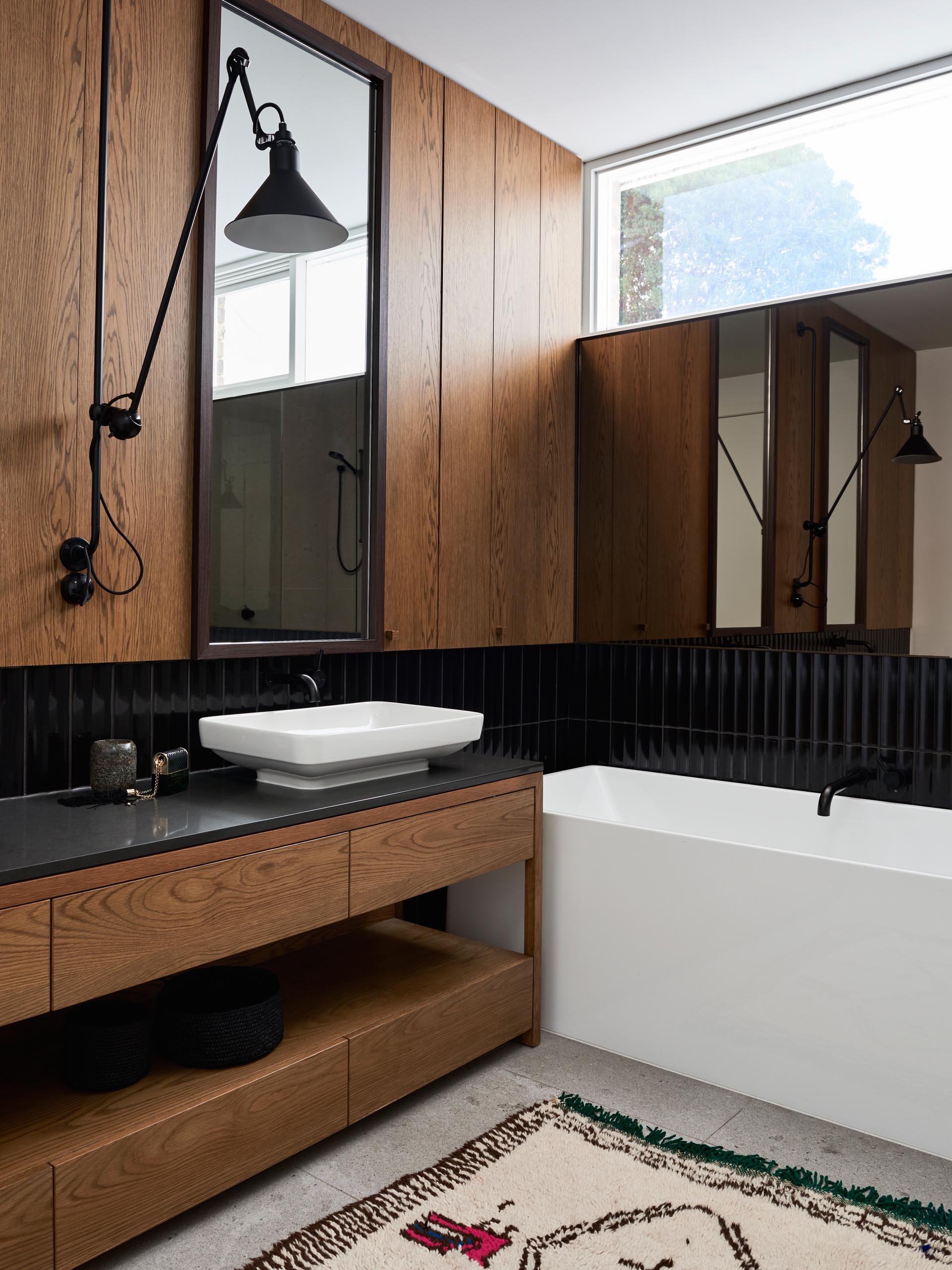 In this modern bathroom, wood paneling and a wood vanity have been paired with black tiles, a white vanity, and white bathtub.