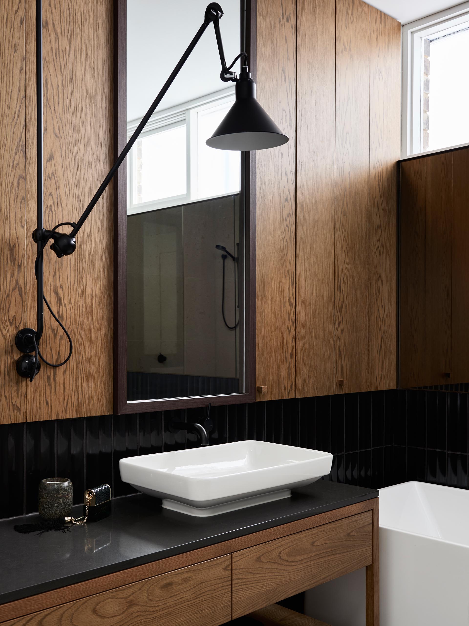 In this modern bathroom, wood paneling and a wood vanity have been paired with black tiles, a white vanity, and white bathtub.