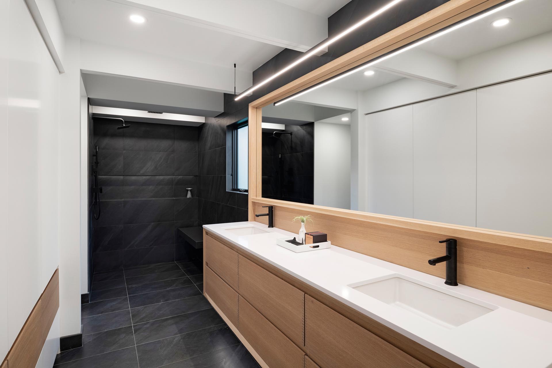 In this mdoern master bathroom, a long wood vanity is topped with a white counter and undermount sinks, while black tile has been used for flooring and the shower.